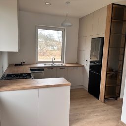 Meble Rostkowski - Jasna kuchnia z drewnianymi blatami i czarną lodówką, okno z widokiem na drzewa w tle.