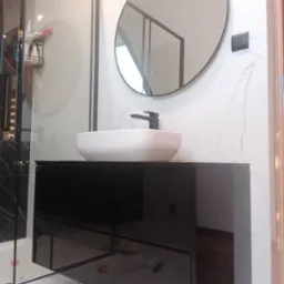 Nowoczesna łazienka z czarną szafką pod umywalkę, okrągłym lustrem i białymi marmurowymi płytkami na ścianie. Widoczna kabina prysznicowa ze szklanymi drzwiami.