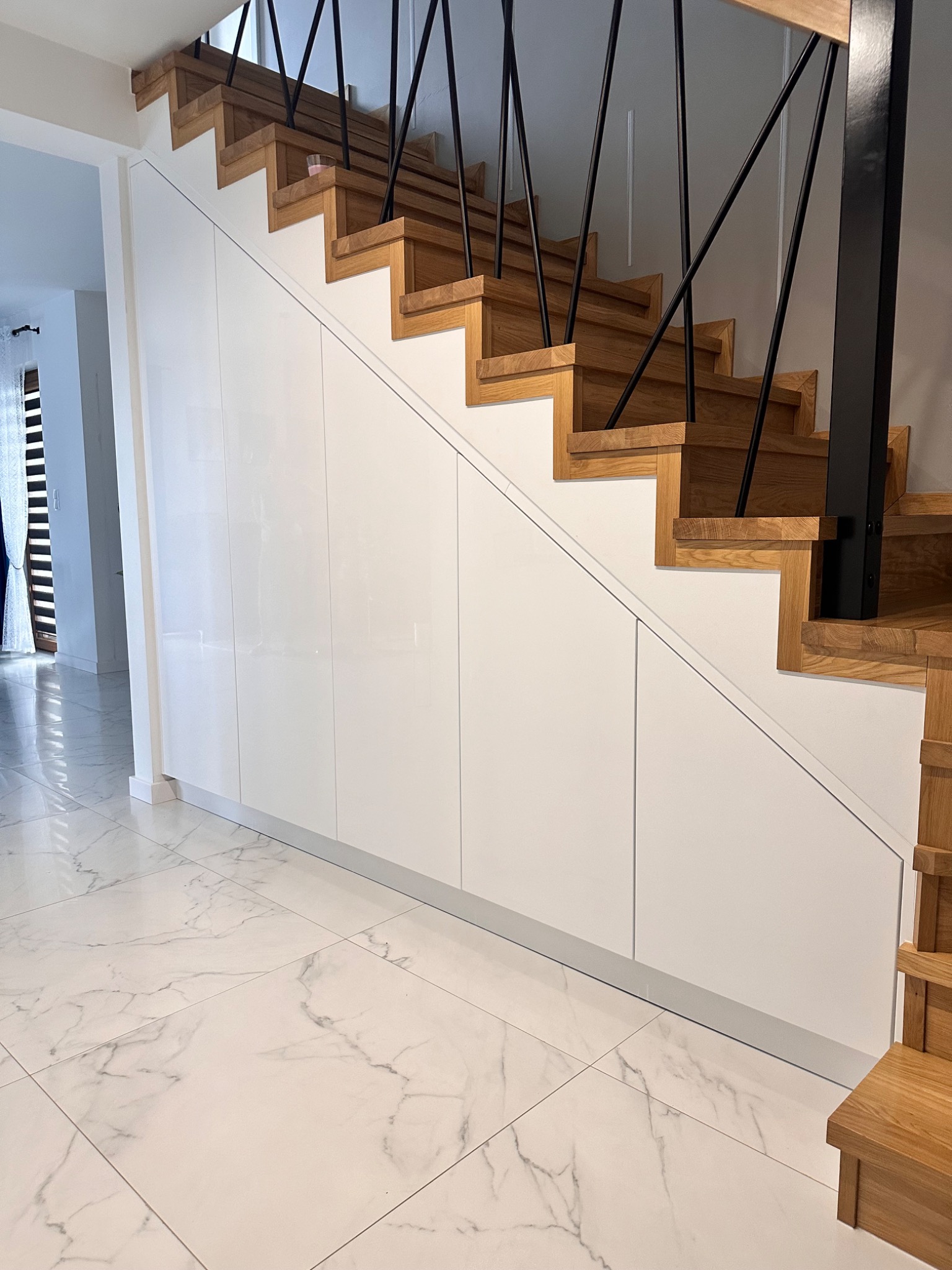 Schody drewniane z czarną balustradą nad zabudową z białymi, lakierowanymi frontami w przedpokoju z marmurową podłogą.