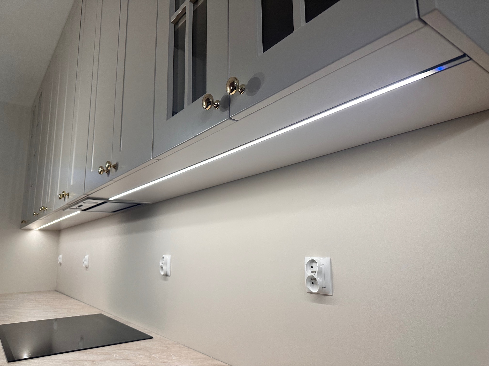 Szare szafki kuchenne z oświetleniem LED podszafkowym i złotymi uchwytami, widoczne gniazdka elektryczne na ścianie i płyta indukcyjna.