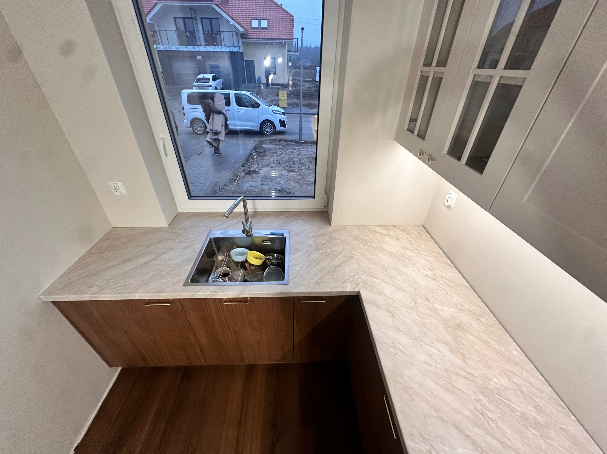 Fragment kuchni z drewnianymi szafkami, blatem imitującym marmur i zlewozmywakiem pełnym naczyń; widok z okna na budowę i samochód dostawczy.