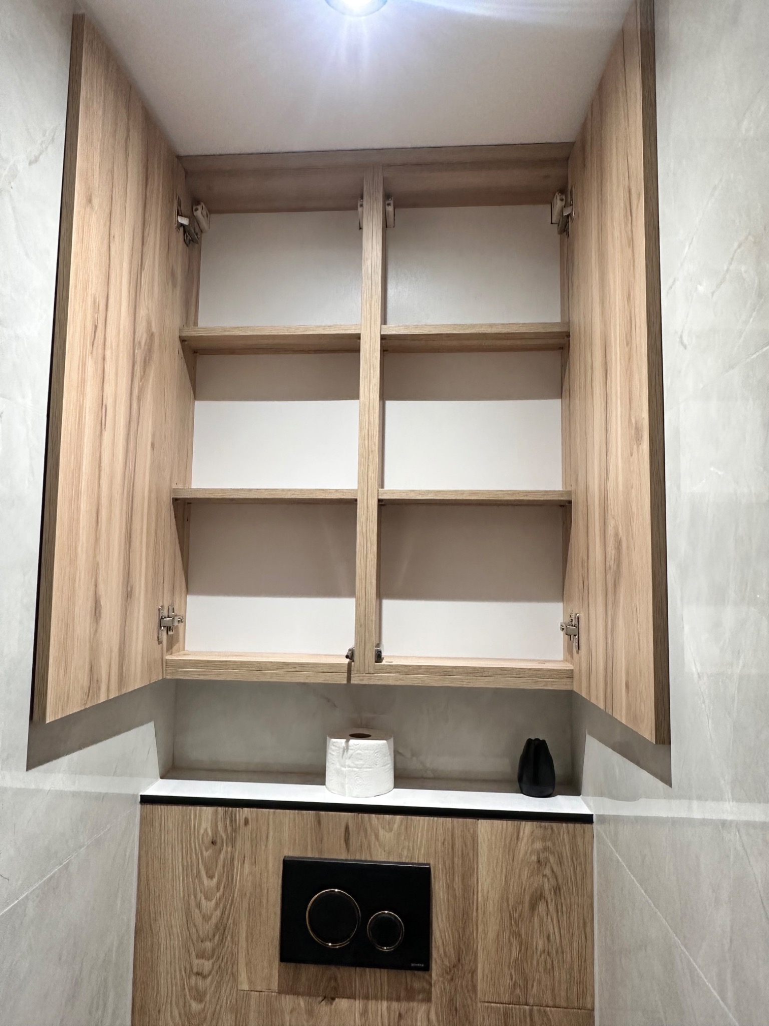 Otwarta szafka łazienkowa z drewnopodobnymi frontami i półkami, z rolką papieru toaletowego i czarnym wazonem na blacie, na tle marmurowej ściany.