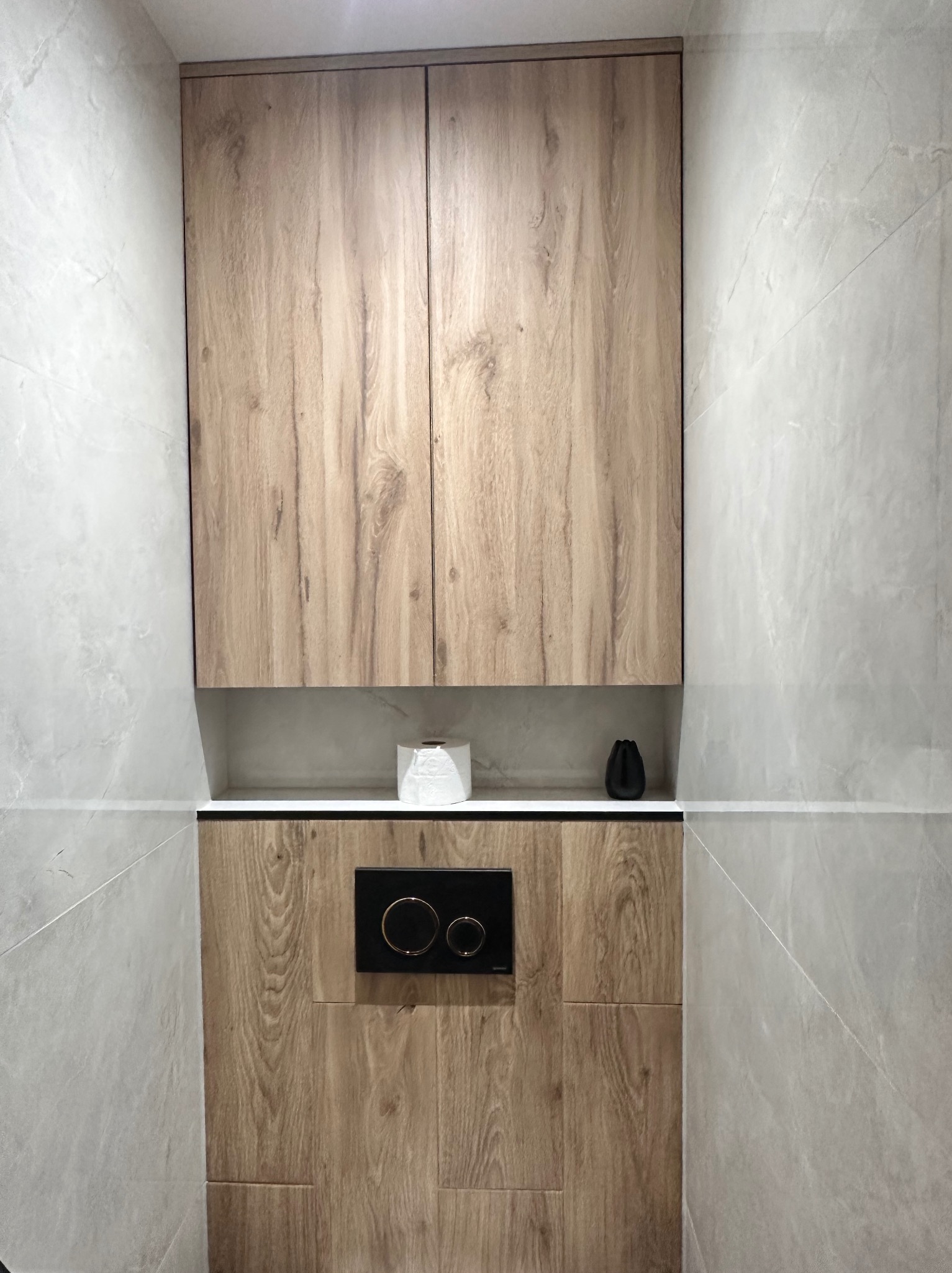 Drewniana szafka wisząca nad zabudowaną toaletą w łazience wykończonej szarymi płytkami imitującymi kamień.