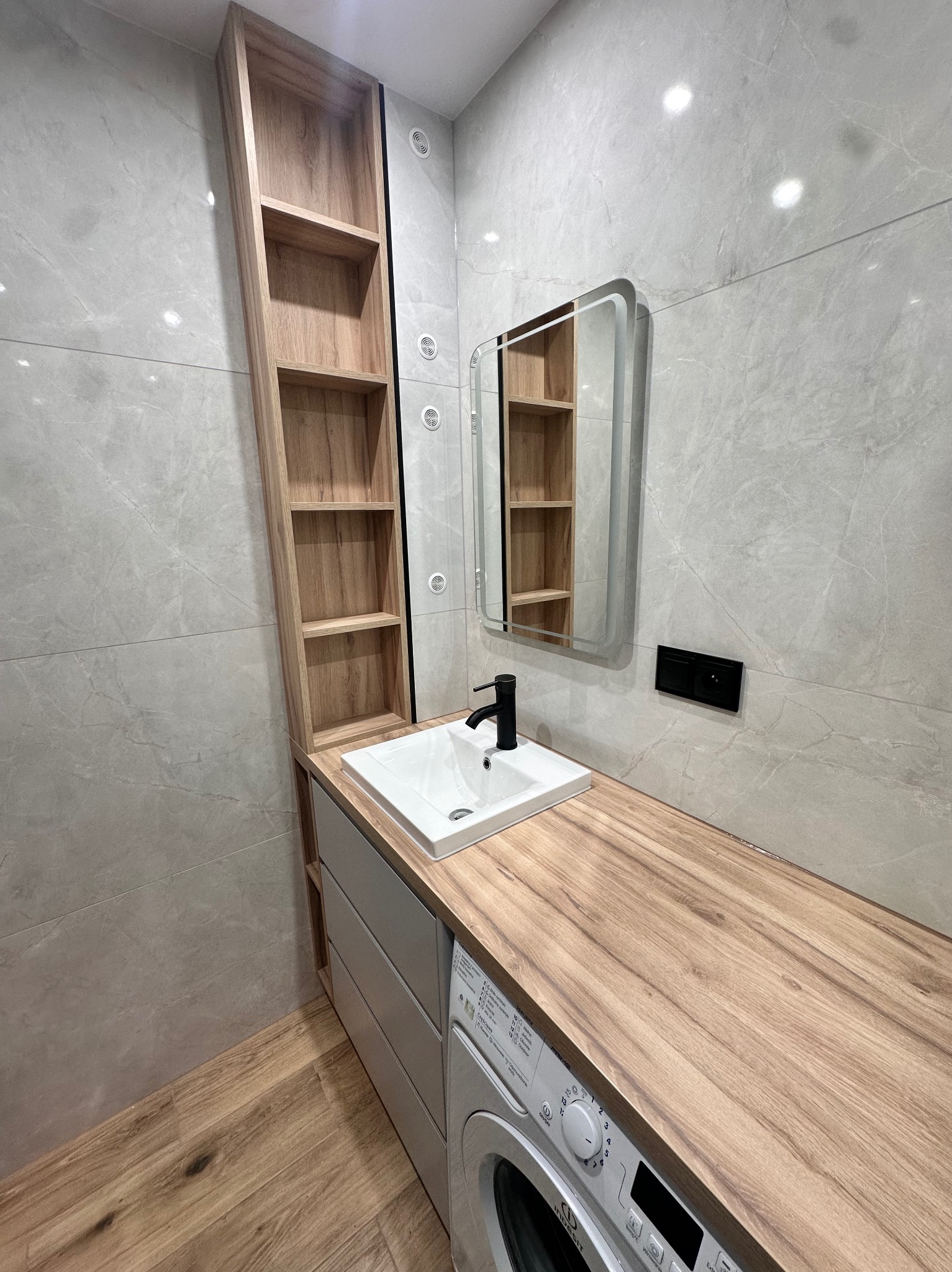 Nowoczesna łazienka z drewnianym blatem nad pralką, białą umywalką, czarną baterią i wysoką, drewnianą półką w zabudowie, uzupełniona o lustro z podświetleniem.