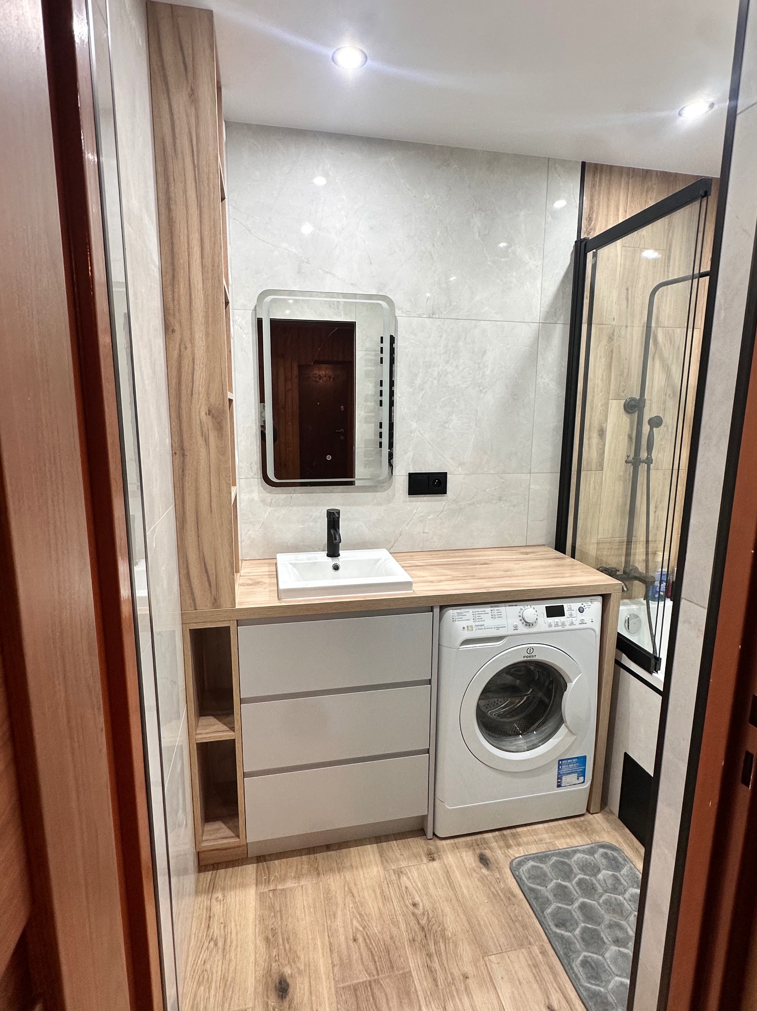 Nowoczesna łazienka w Małkini z zabudową meblową na wymiar, obejmującą szafki z szufladami, umywalkę nablatową, pralkę oraz lustro z podświetleniem, wszystko w jasnej kolorystyce z drewnianymi...