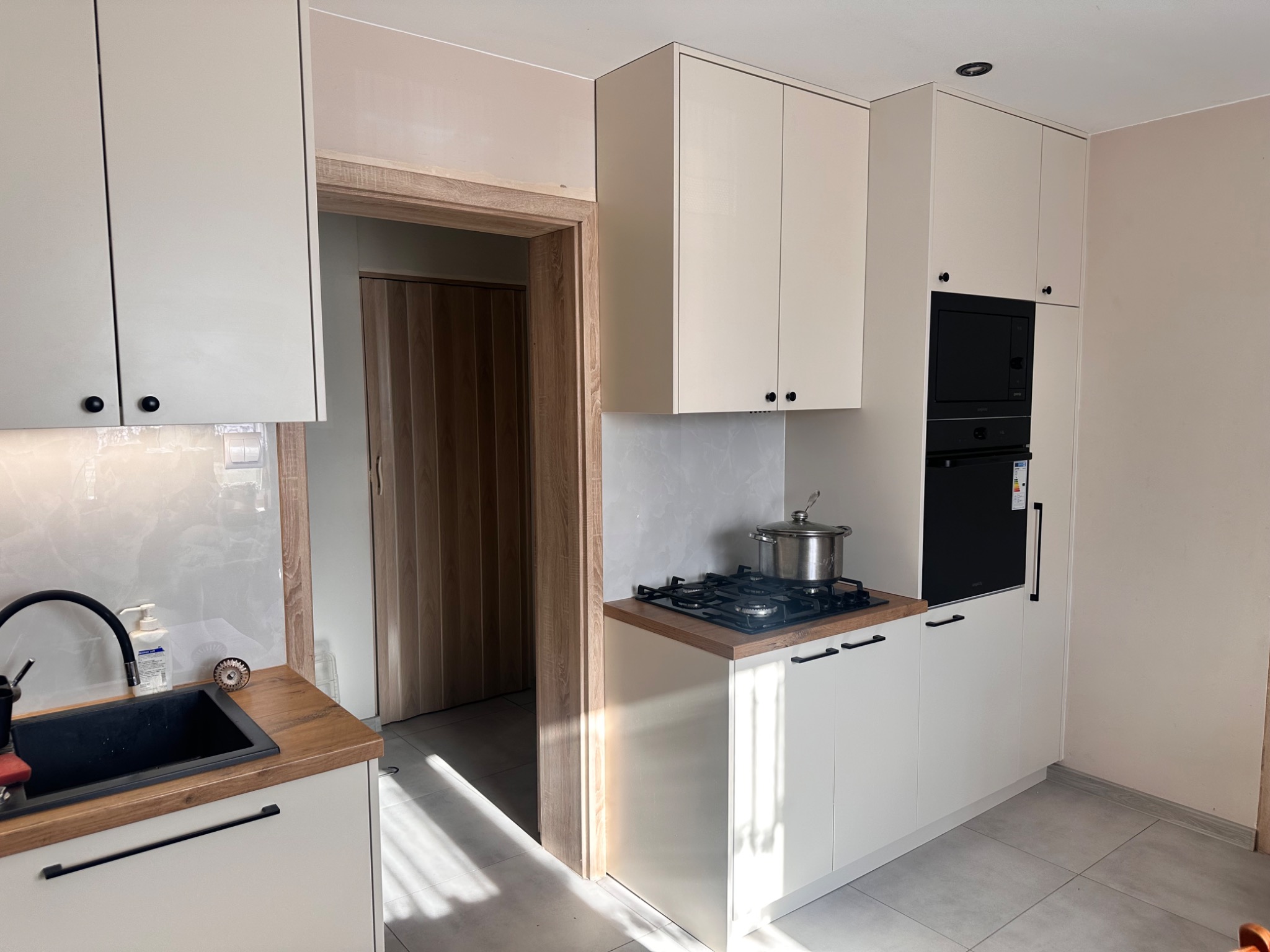 Jasna kuchnia z drewnianym blatem, czarnym zlewozmywakiem i piekarnikiem, widoczny garnek na płycie grzewczej i drzwi z drewnianą ościeżnicą.