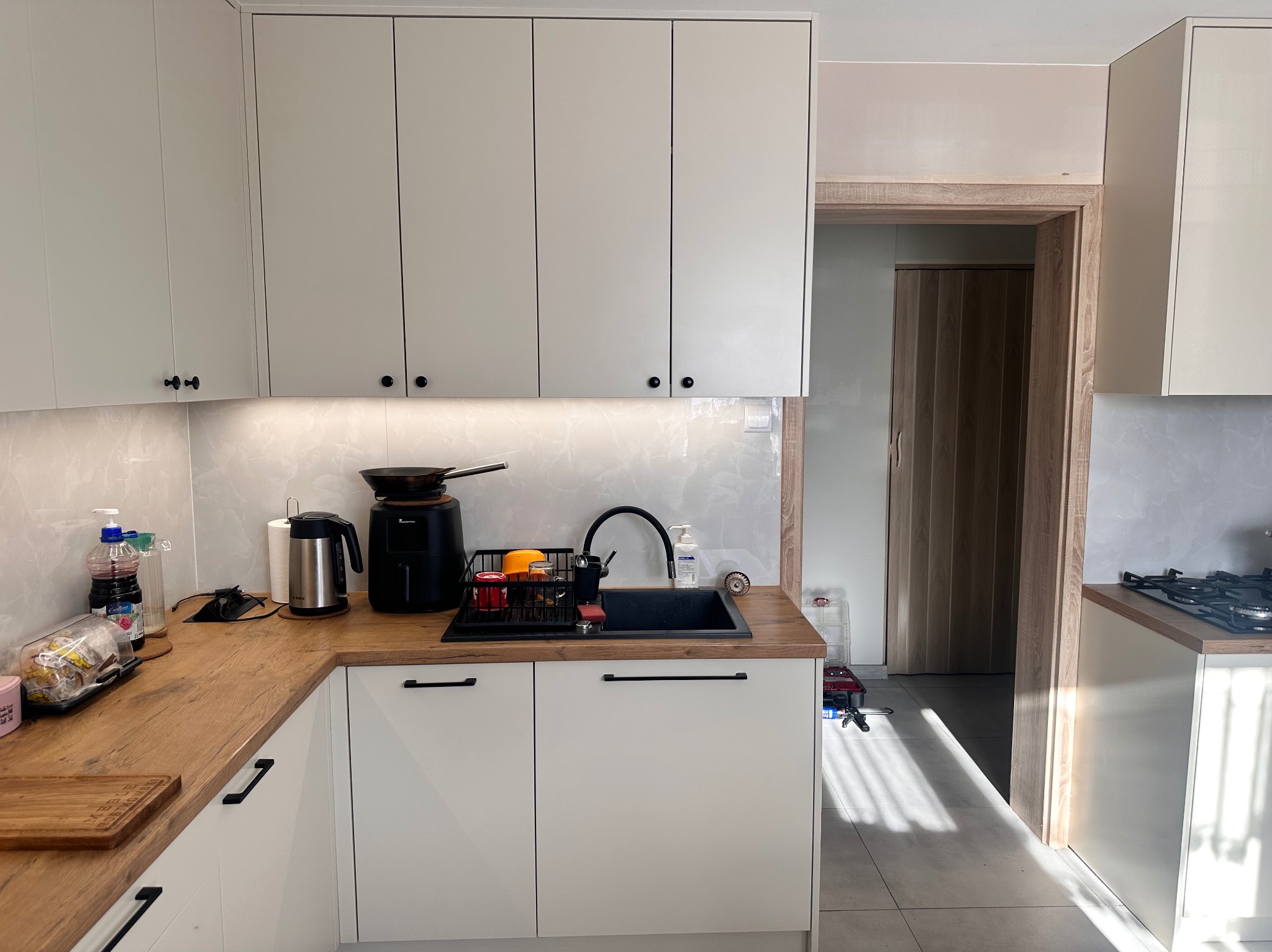 Jasna kuchnia z drewnianym blatem, czarnym zlewem i suszarką na naczynia. Widoczne sprzęty AGD, takie jak czajnik i frytkownica beztłuszczowa. W tle drzwi harmonijkowe.