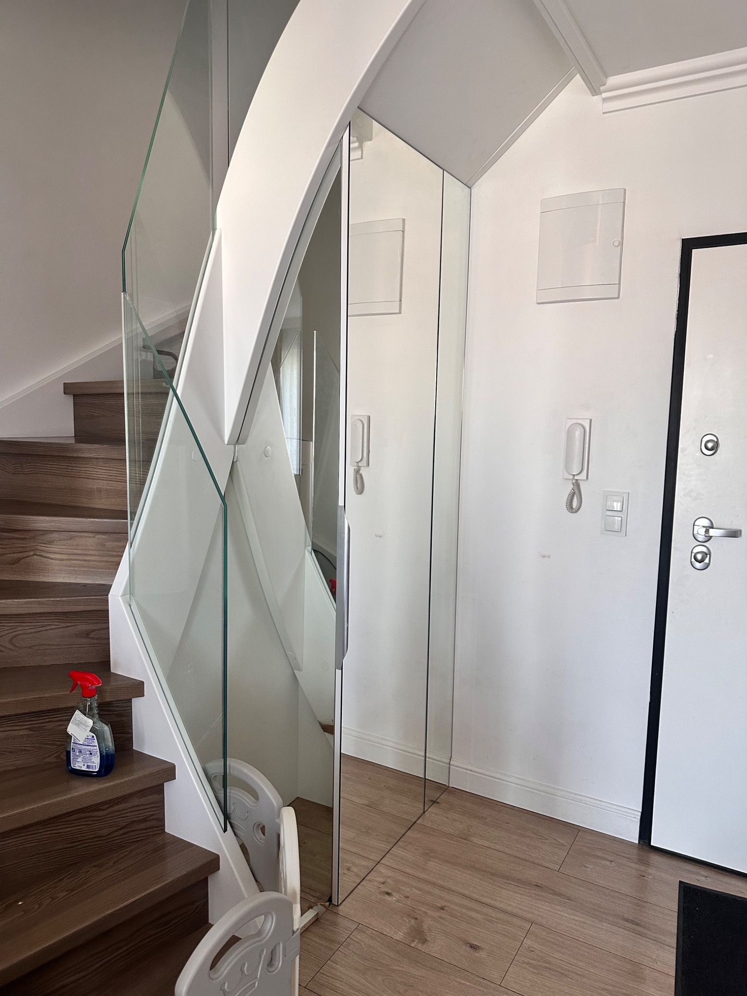 Nowoczesne schody z drewnianymi stopniami i szklaną balustradą, przylegające do lustrzanej szafy w przedpokoju z drewnianą podłogą i białą ścianą z domofonem.