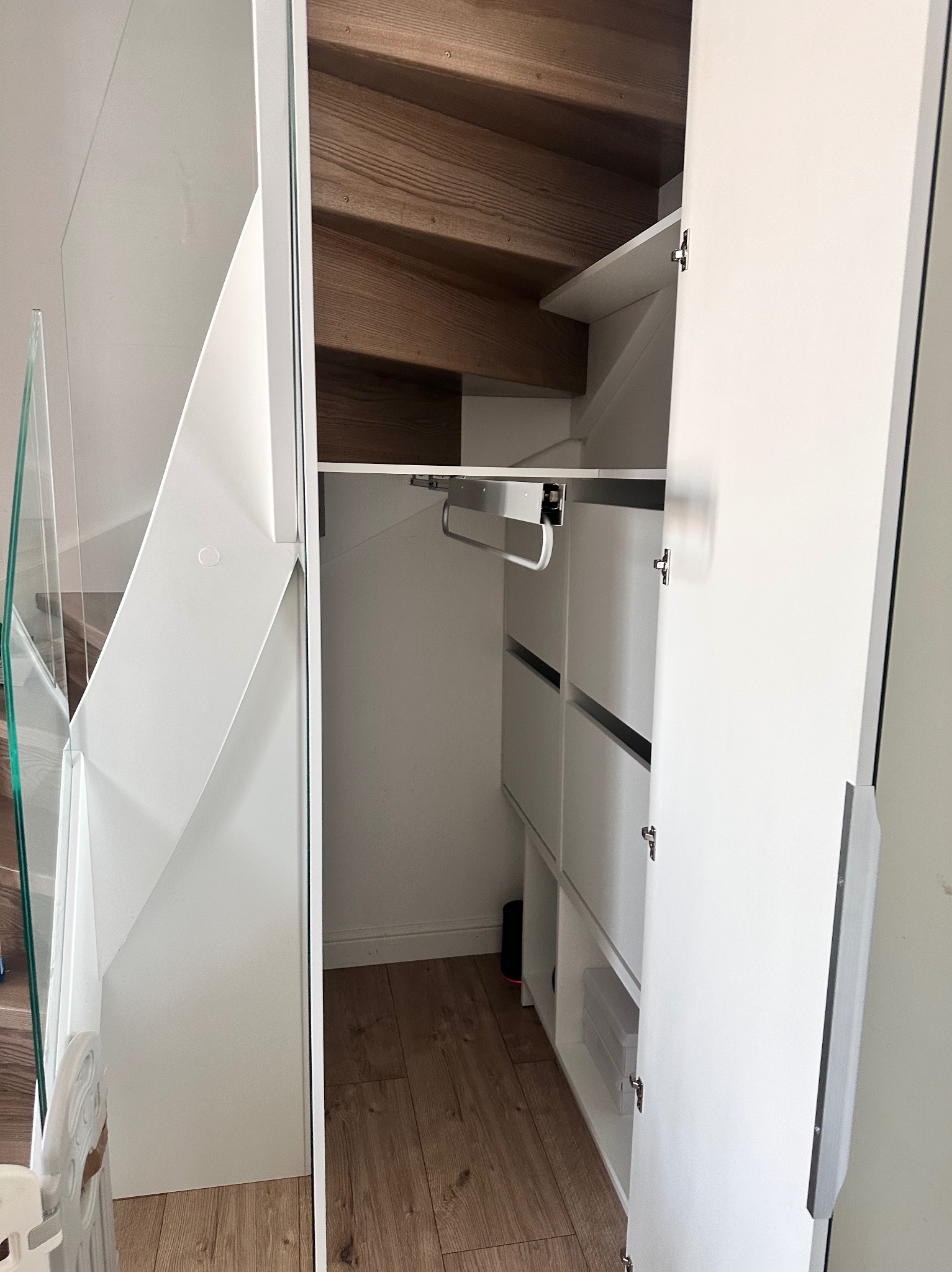 Biała szafa wnękowa pod schodami z szufladami i drążkiem na ubrania, widoczna od strony otwartych drzwi, z elementami szklanymi.