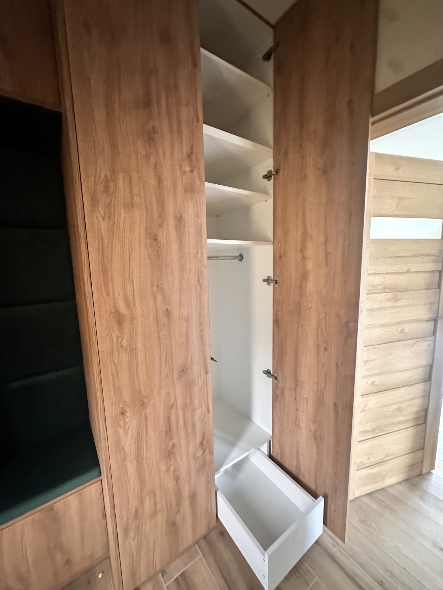 Wnętrze szafy wnękowej z drewnianymi frontami, widoczna półka, drążek i wysuwana szuflada, obok zielone siedzisko tapicerowane.