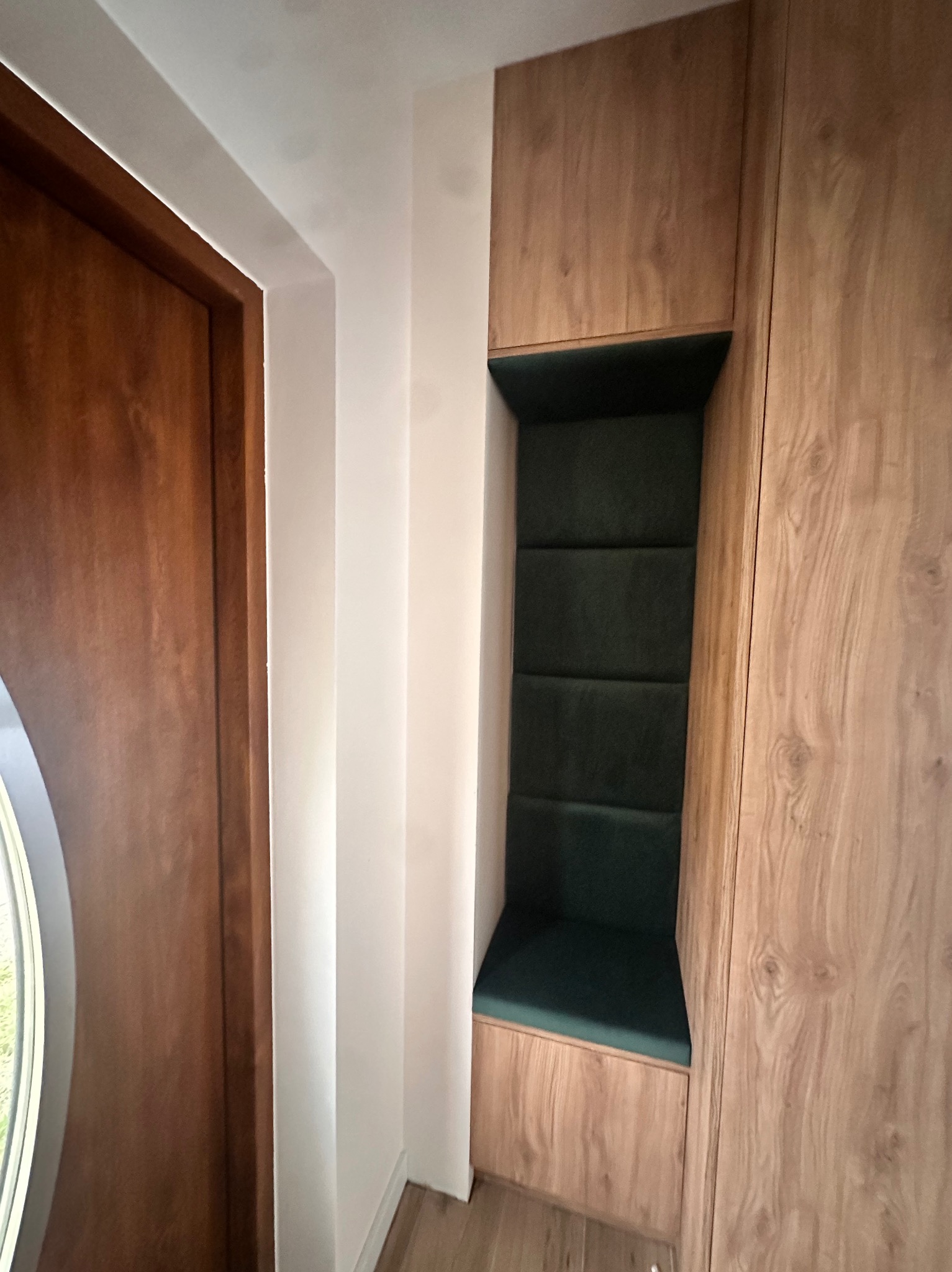 Wnęka z siedziskiem i tapicerowanym panelem ściennym w kolorze butelkowej zieleni, obudowana płytą meblową w odcieniach drewna, umieszczona obok drewnianych drzwi z okrągłym przeszkleniem.