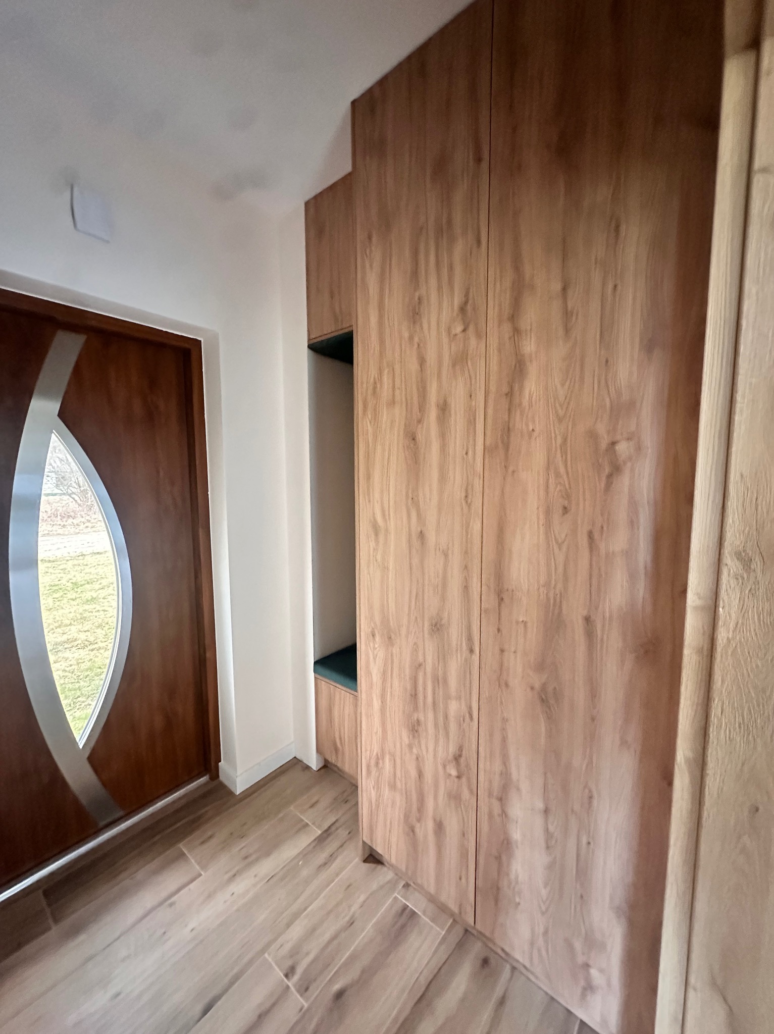 Drewniana szafa wnękowa z siedziskiem w przedpokoju, widoczna obok drzwi wejściowych z dekoracyjnym przeszkleniem w domu w Małkini.