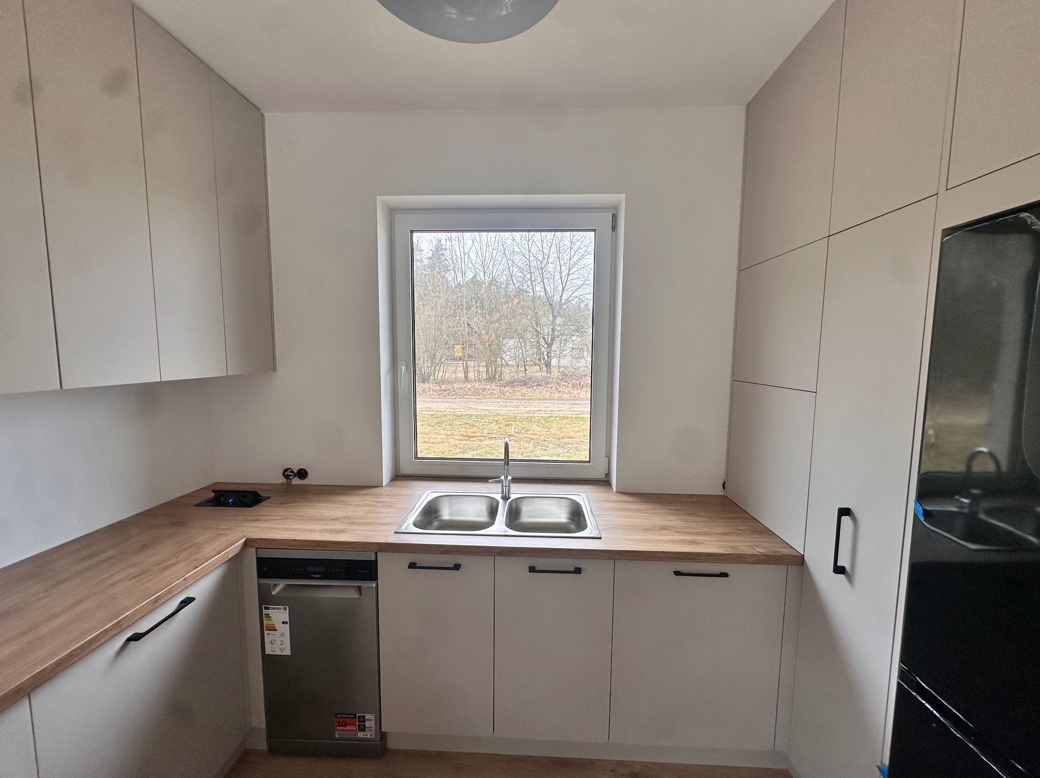 Jasna kuchnia z drewnianym blatem, zlewozmywakiem dwukomorowym pod oknem i zabudowaną zmywarką, widok na drzewa za oknem.