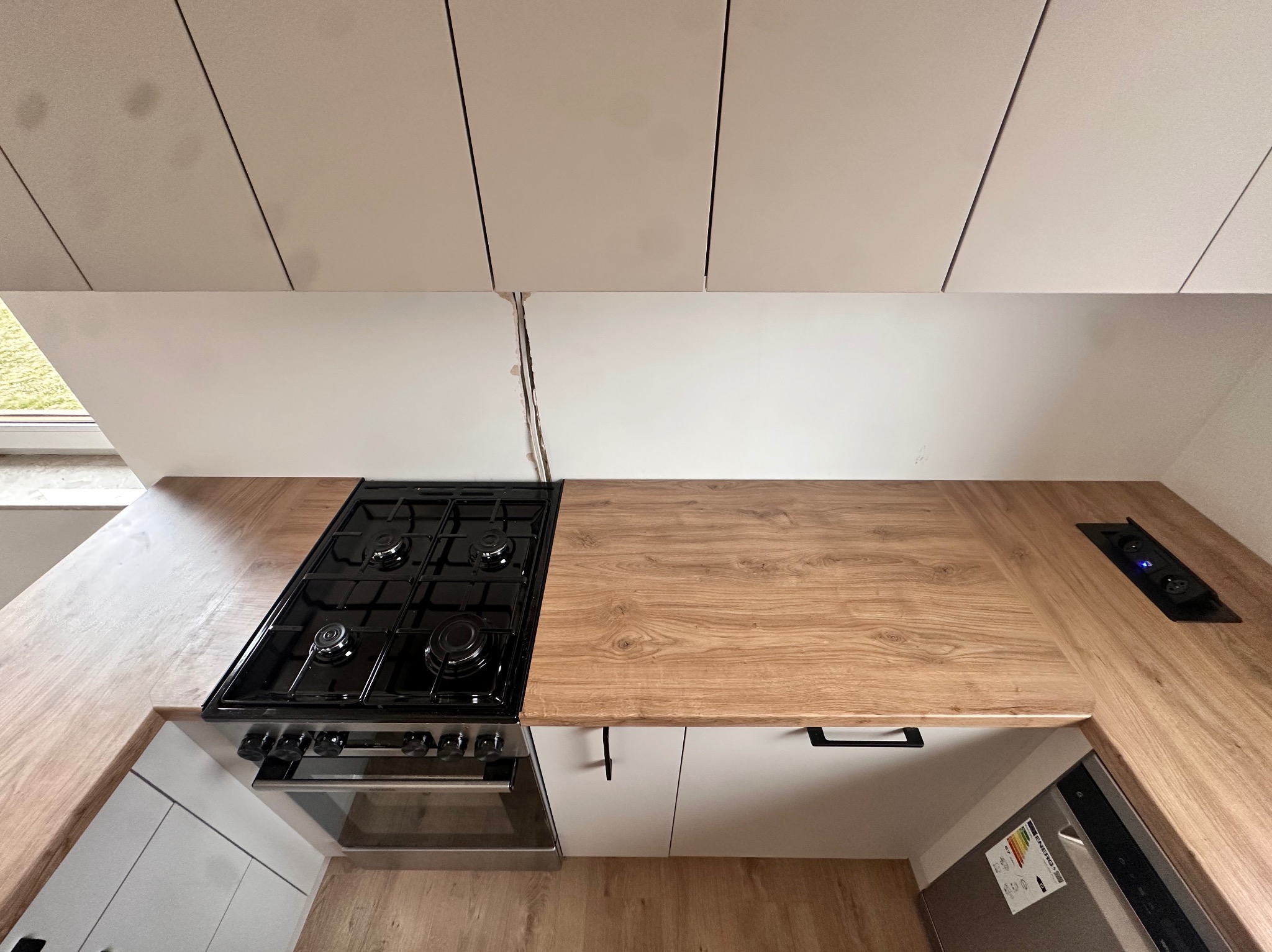 Nowoczesna kuchnia z blatem drewnianym, czarną płytą gazową i białymi szafkami, widok z góry na ukośnym poddaszu, widoczne gniazdka w blacie