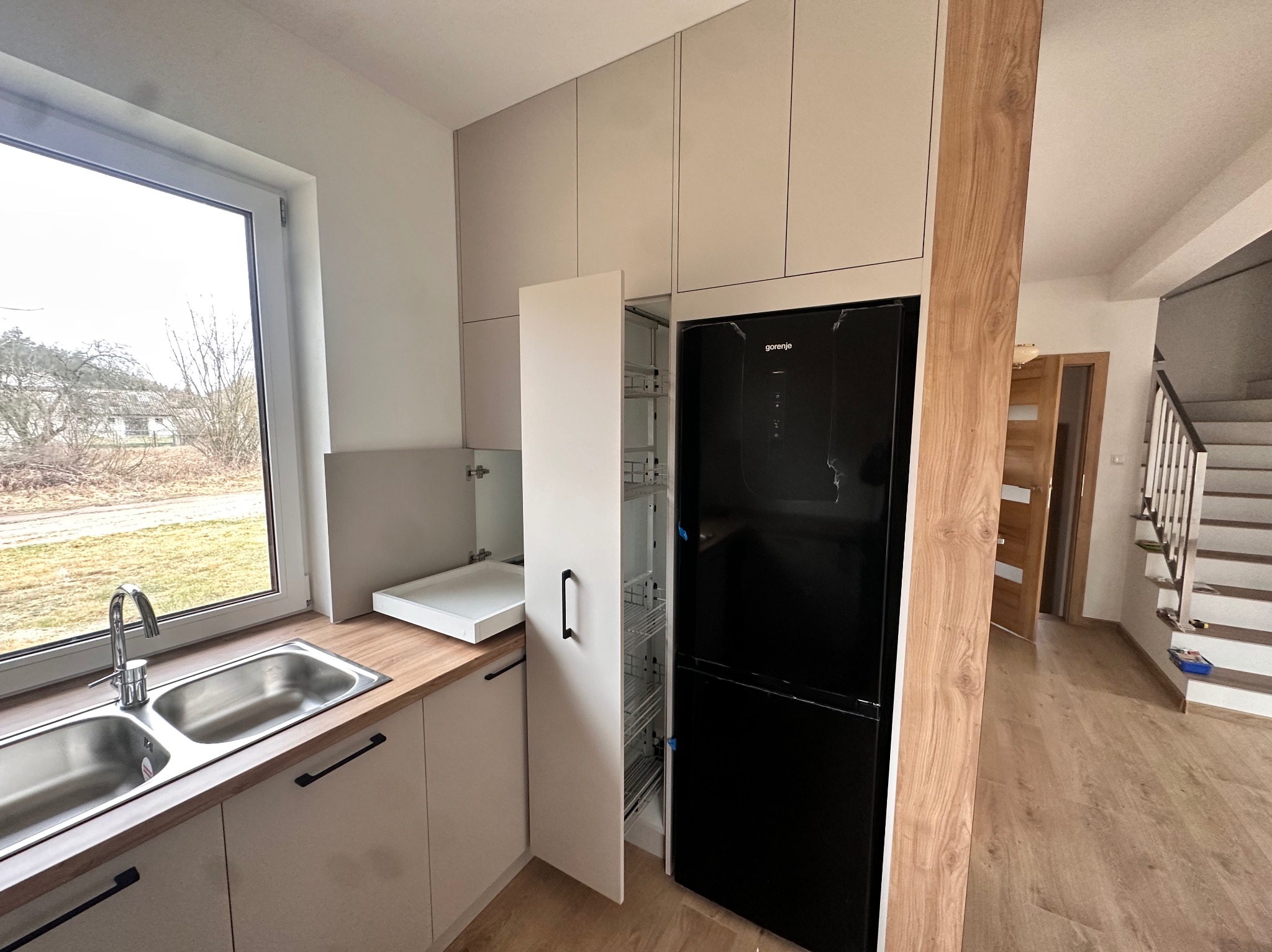 Jasna kuchnia z drewnianym blatem, stalowym zlewem i otwartą szafką cargo obok czarnej lodówki, widok na schody w tle.