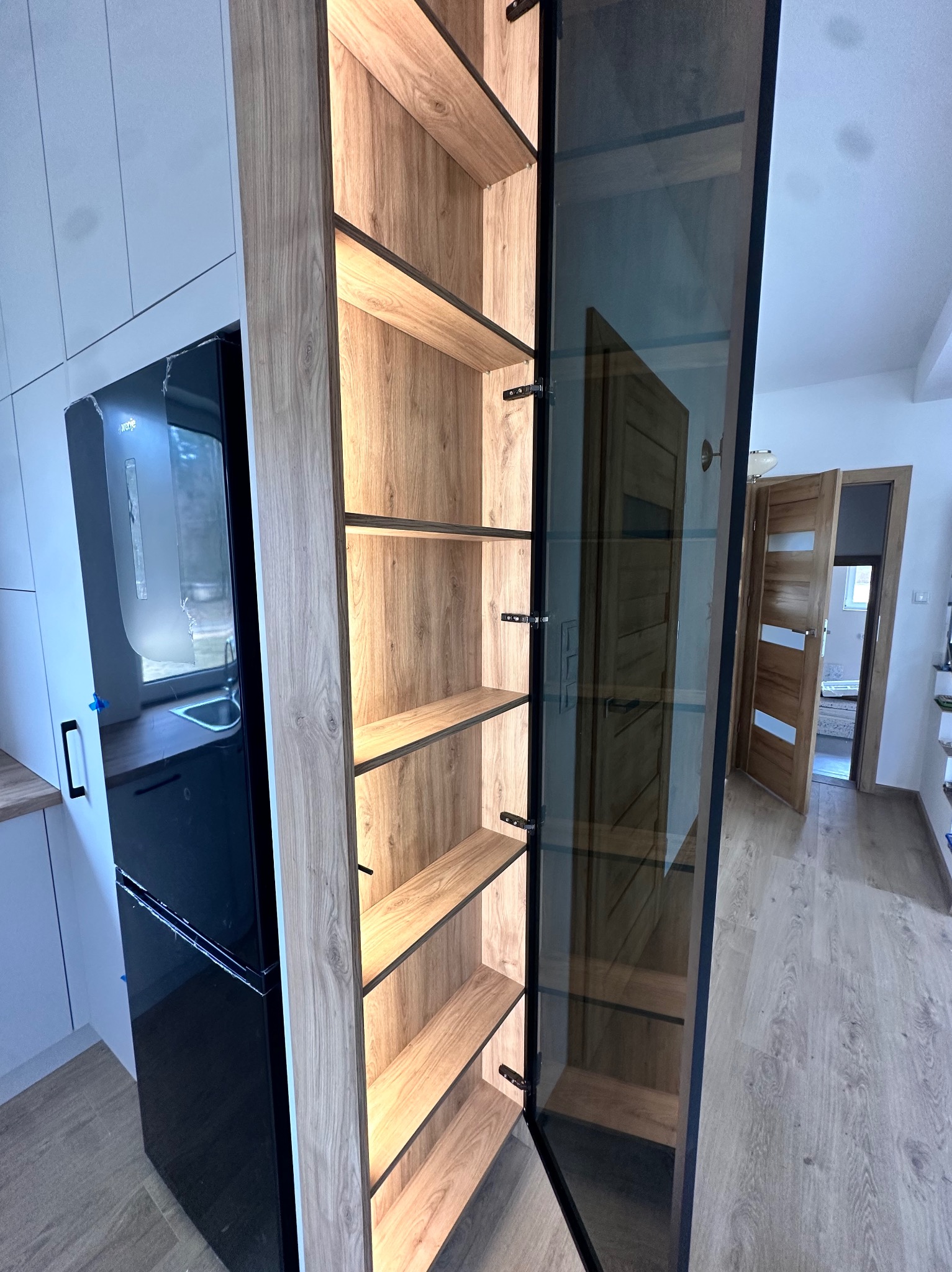 Wysoka szafa wnękowa z oświetlonymi półkami z drewna, przeszklone drzwiczki z czarną ramą, widok na wnętrze domu.