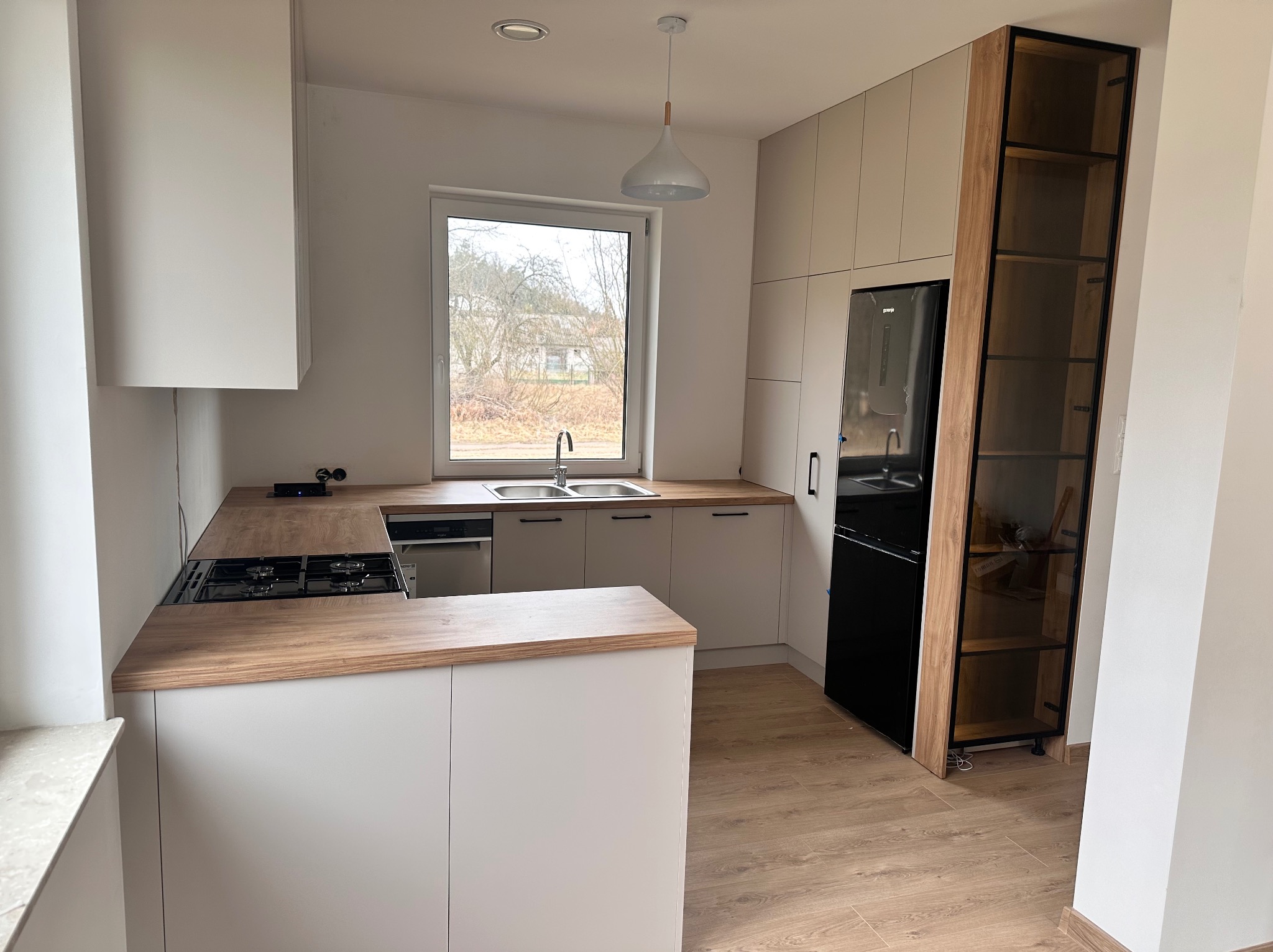 Jasna kuchnia z drewnianymi blatami i czarną lodówką, okno z widokiem na drzewa w tle.