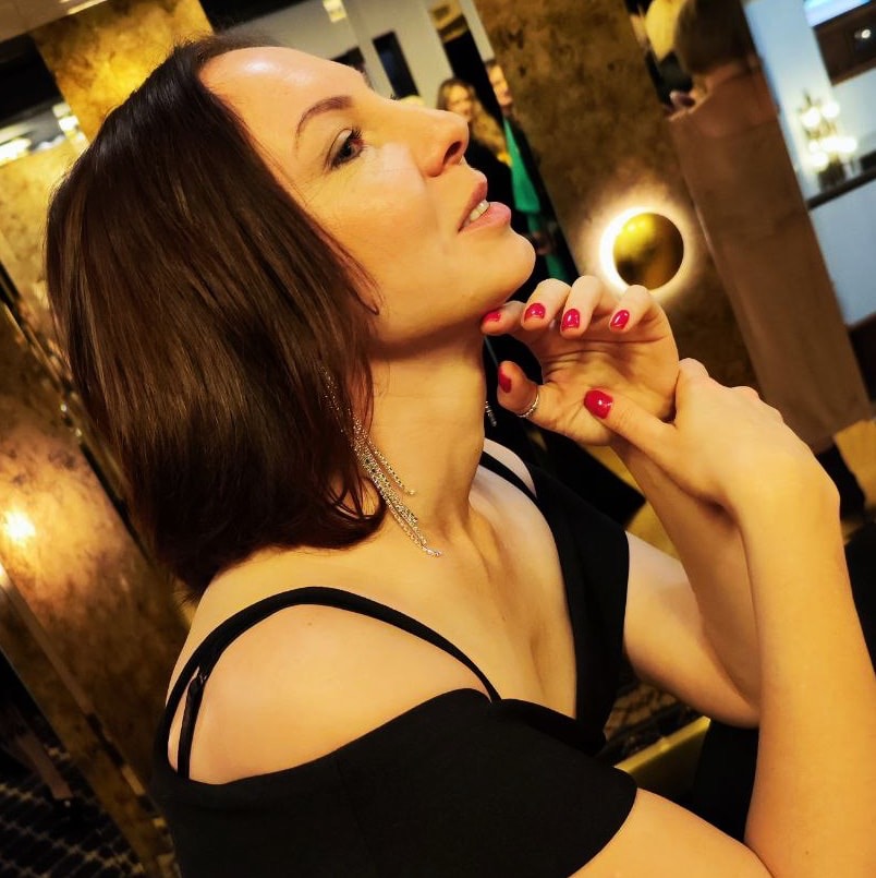 Kobieta z ciemnymi włosami, w czarnej sukience z odkrytymi ramionami i długimi, srebrnymi kolczykami, spogląda w górę w eleganckim wnętrzu z dekoracyjnym oświetleniem.