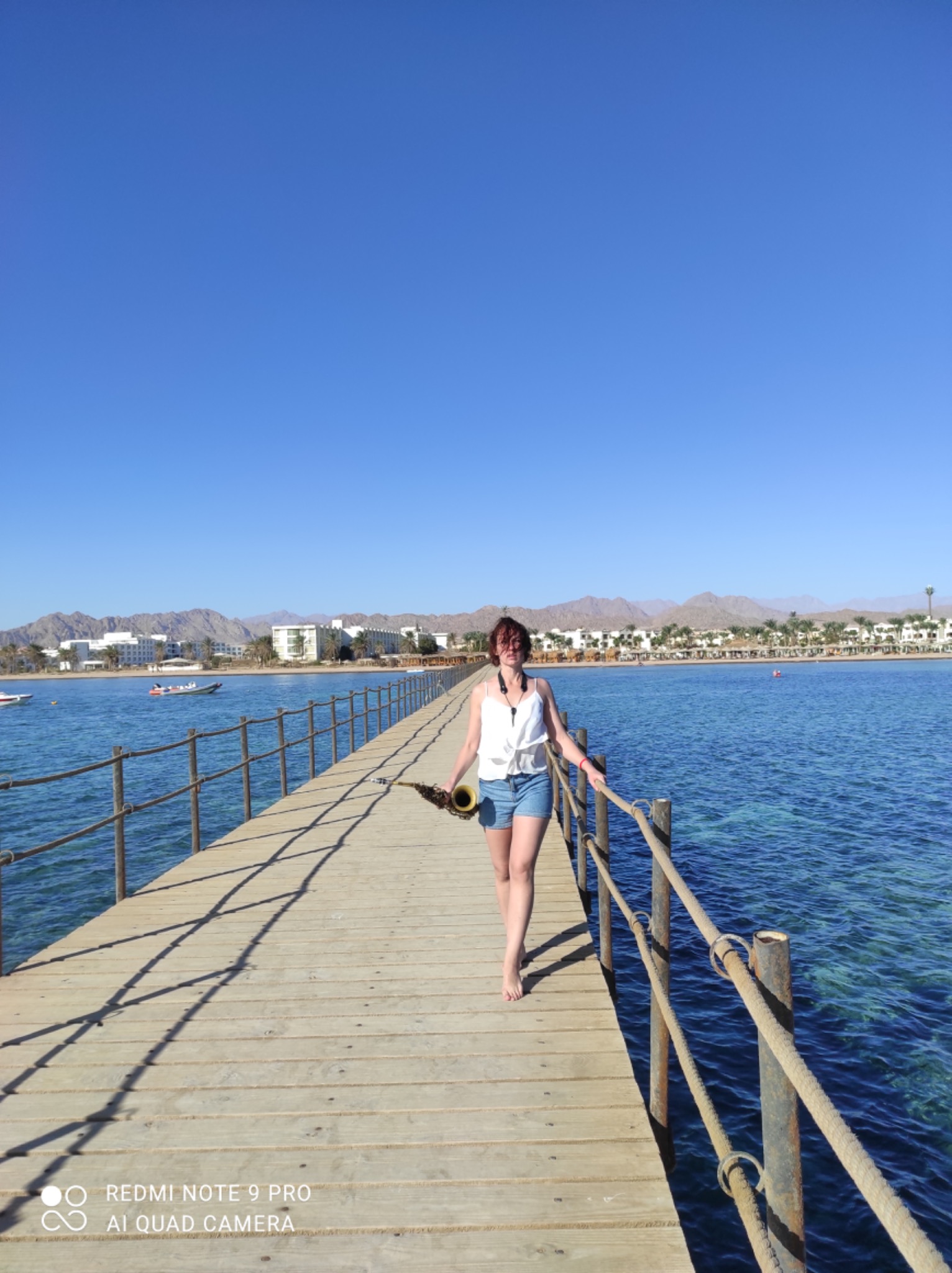 Kobieta idzie boso po drewnianym molo nad morzem, trzymając saksofon w ręce, w tle budynki hotelowe i góry.