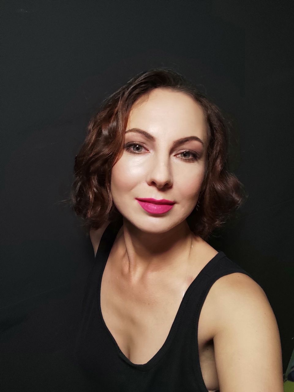 Portret kobiety z krótkimi, brązowymi, kręconymi włosami i różową szminką na tle ciemnoszarego tła.