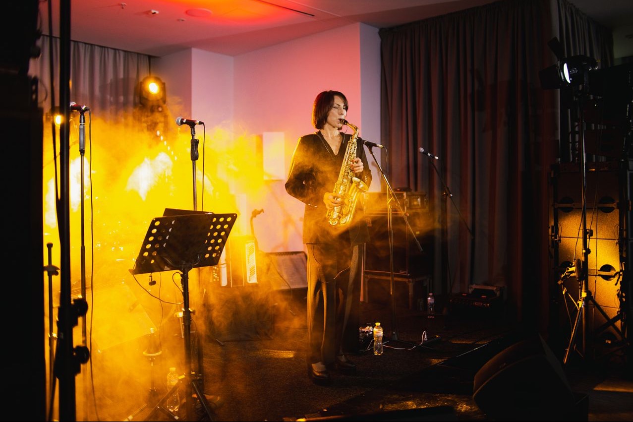 Saksofonistka w czarnym stroju gra na saksofonie na scenie w oświetleniu scenicznym, widoczny statyw na nuty i mikrofony.