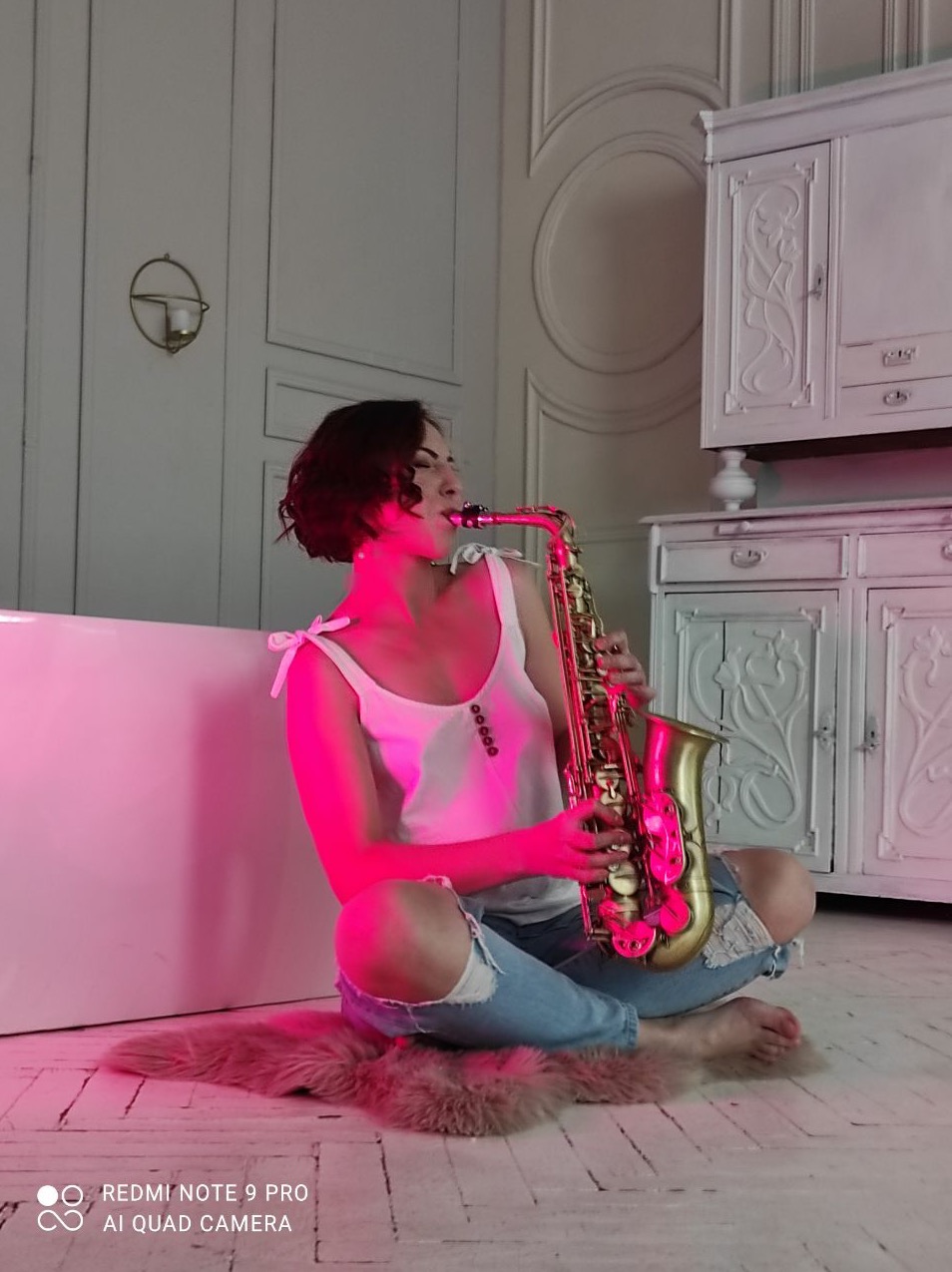 Kobieta siedzi na podłodze, grając na saksofonie w pokoju z białymi meblami i różowym oświetleniem.