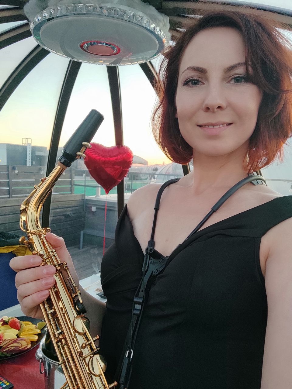 Uśmiechnięta kobieta w czarnej sukience trzymająca złoty saksofon na tle zachodzącego słońca, czerwony balon w kształcie serca i talerz owoców na stole.