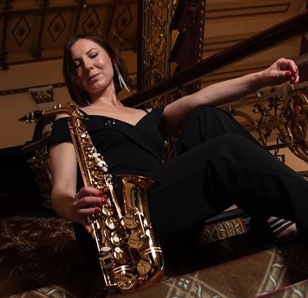 Kobieta w czarnym kombinezonie siedzi na schodach z pozłacaną balustradą, trzymając saksofon tenorowy w dłoni.