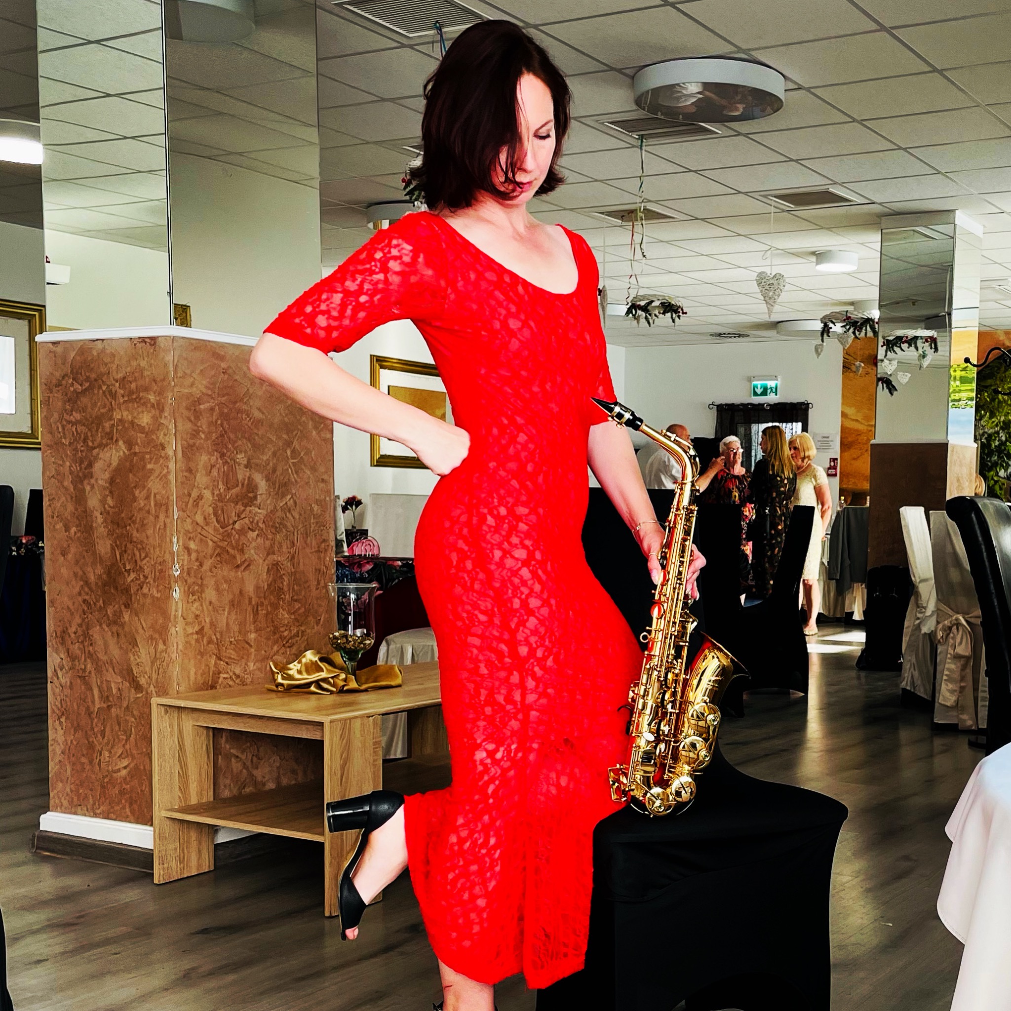 Kobieta w czerwonej, koronkowej sukience pozuje z saksofonem w sali bankietowej, z widocznymi stolikami i gośćmi w tle.