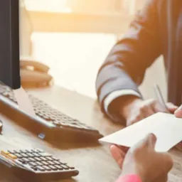 Mężczyzna w garniturze wręcza dokument drugiej osobie przy biurku z monitorem i kalkulatorem.