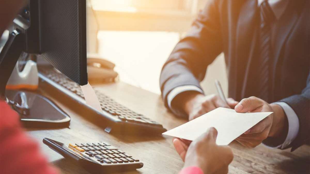 Mężczyzna w garniturze wręcza dokument drugiej osobie przy biurku z monitorem i kalkulatorem.