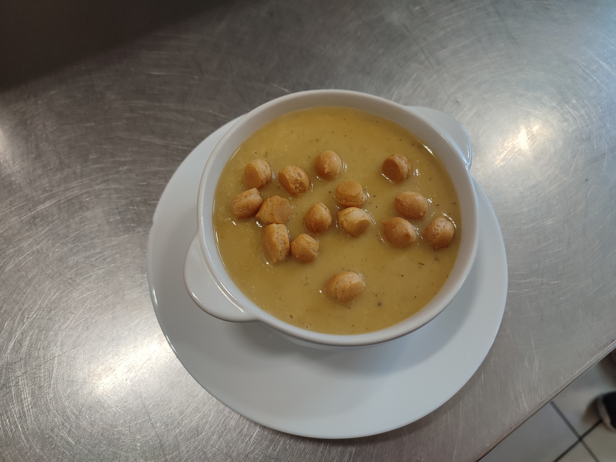 Kremowa zupa z groszkiem ptysiowym w białej miseczce na białym talerzu, ustawiona na metalowym blacie.