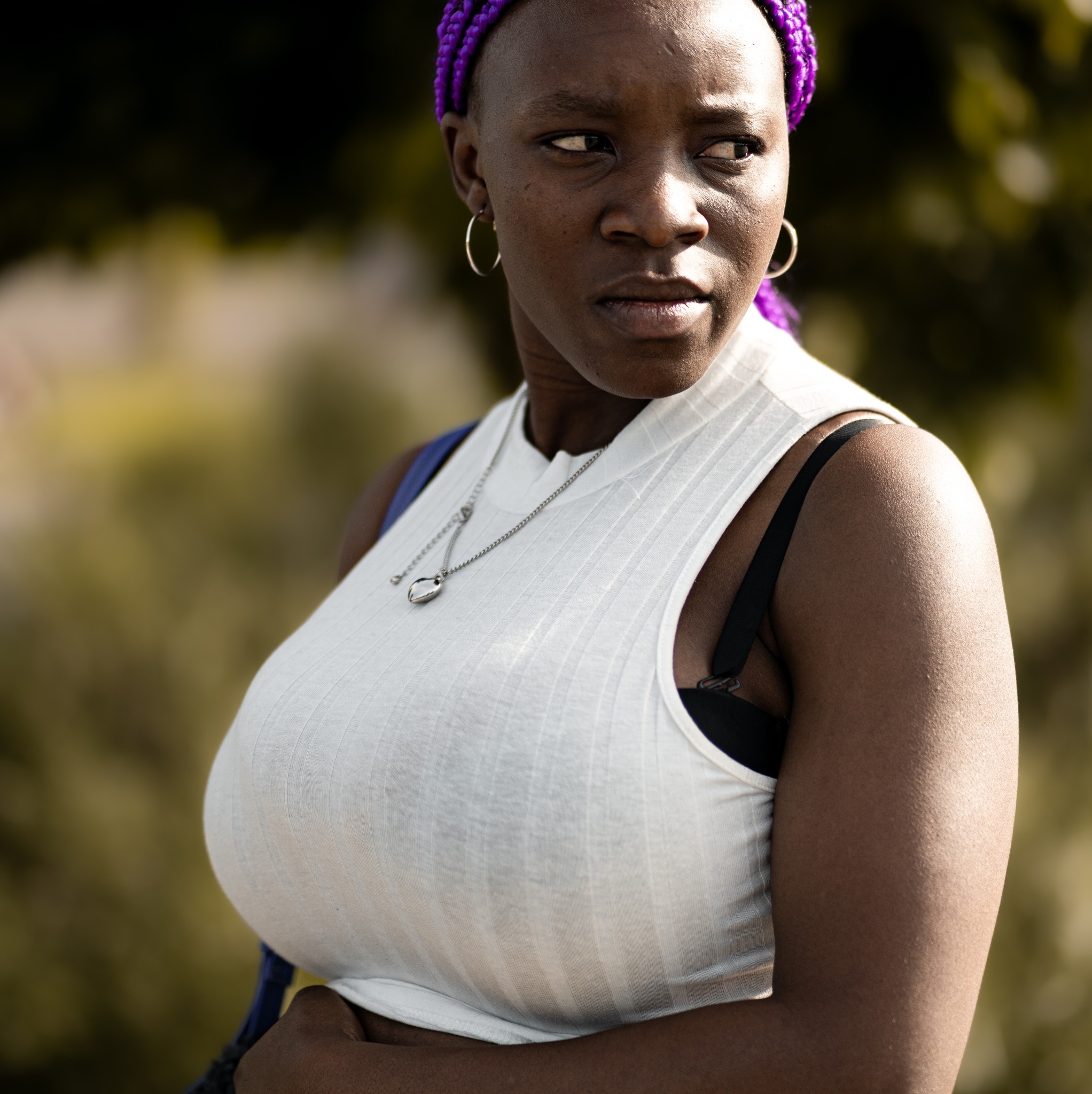 Portret kobiety z fioletowymi warkoczykami, srebrnymi kolczykami i naszyjnikiem, ubrana w biały top na tle zieleni.