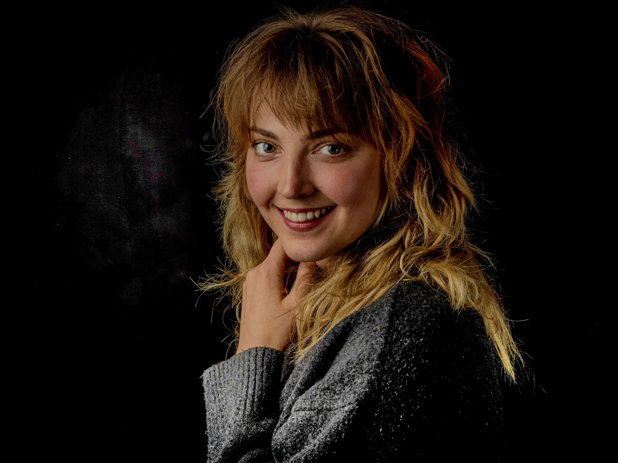 Portret uśmiechniętej kobiety z blond włosami w szarym swetrze na czarnym tle, dłoń pod brodą.