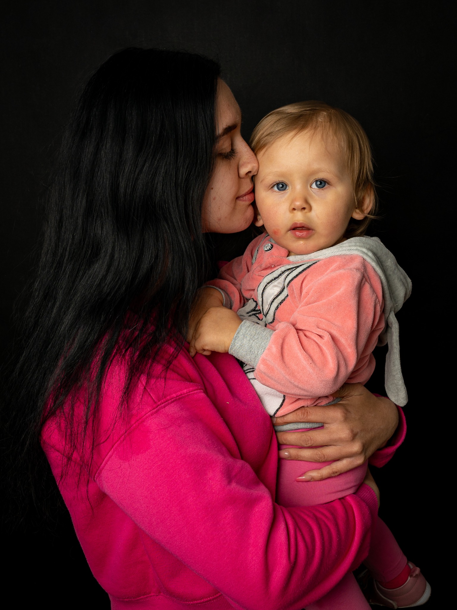Portret matki całującej w czoło roczne dziecko ubrane w różowy welurowy dres z kapturem na czarnym tle.