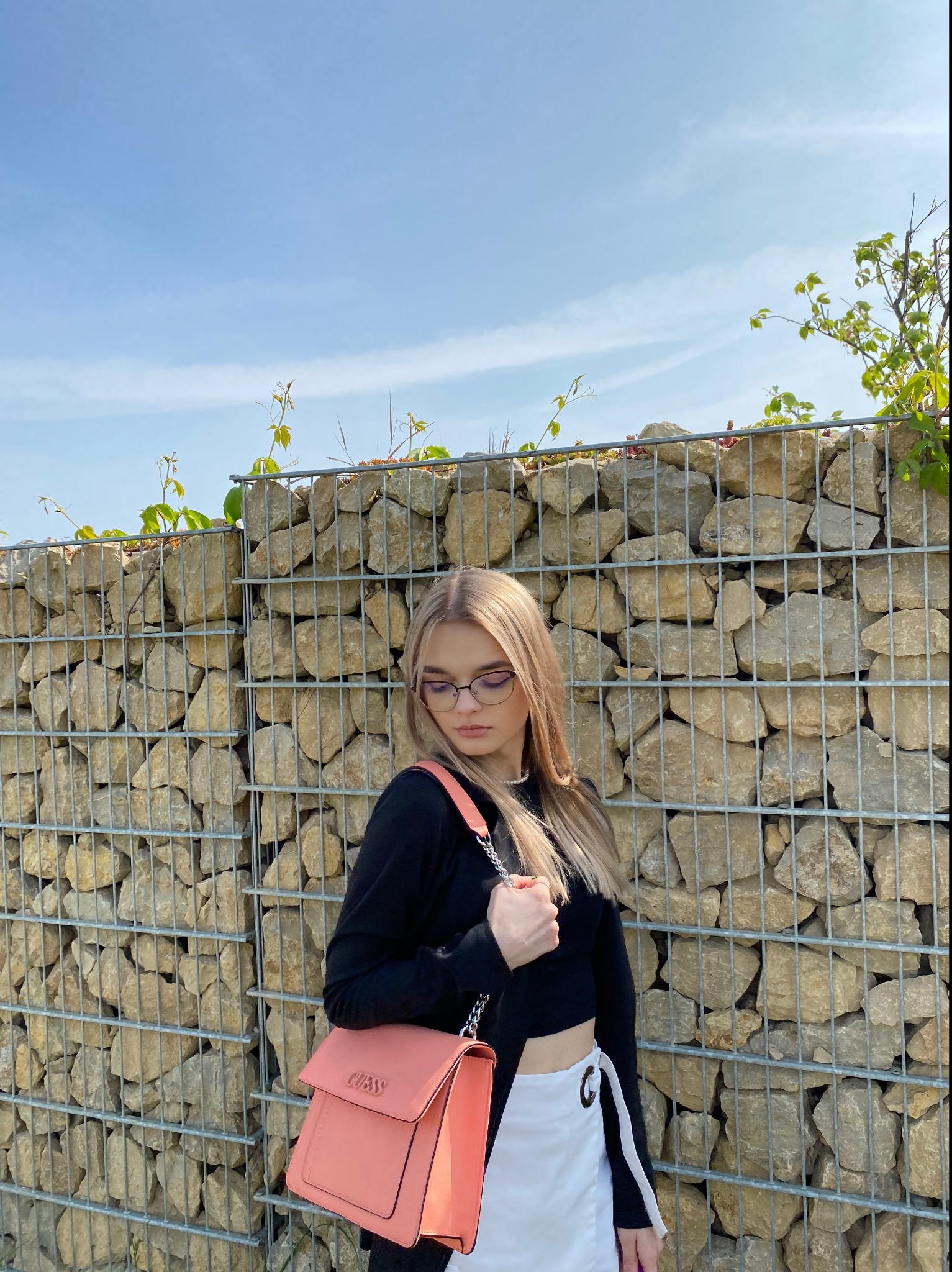 Młoda kobieta w okularach, czarnym topie i białej spódnicy pozuje z różową torebką na tle kamiennej ściany z metalową siatką i pnącymi roślinami.