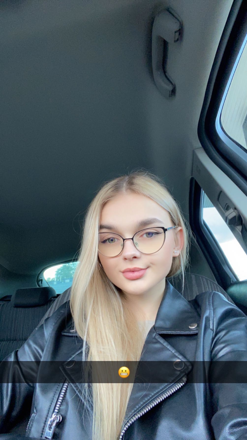 Portret uśmiechniętej blondynki w okularach i czarnej skórzanej kurtce, siedzącej w samochodzie.