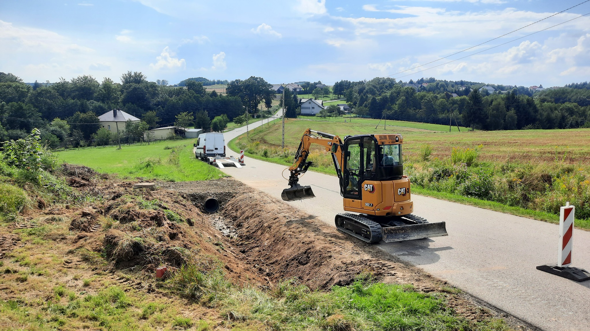 Żółta mini koparka CAT 302.7 stoi na asfaltowej drodze obok wykopu z ułożoną rurą drenażową, w tle biały samochód dostawczy na lawecie i wiejski krajobraz.