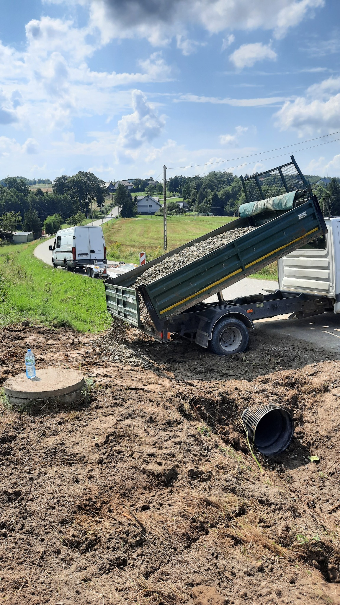 Przyczepa wywrotka z ładunkiem kruszywa wysypuje materiał na plac budowy, widoczny właz studzienki kanalizacyjnej, rura drenażowa i zaparkowany bus z przyczepą w tle.