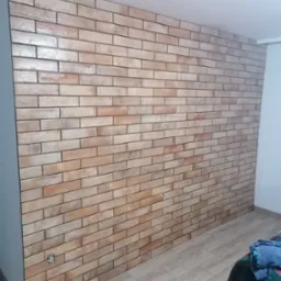 Ściana wyłożona beżowo-brązowymi płytkami imitującymi cegłę, z widocznym gniazdkiem elektrycznym i fragmentem podłogi z paneli w odcieniach drewna.