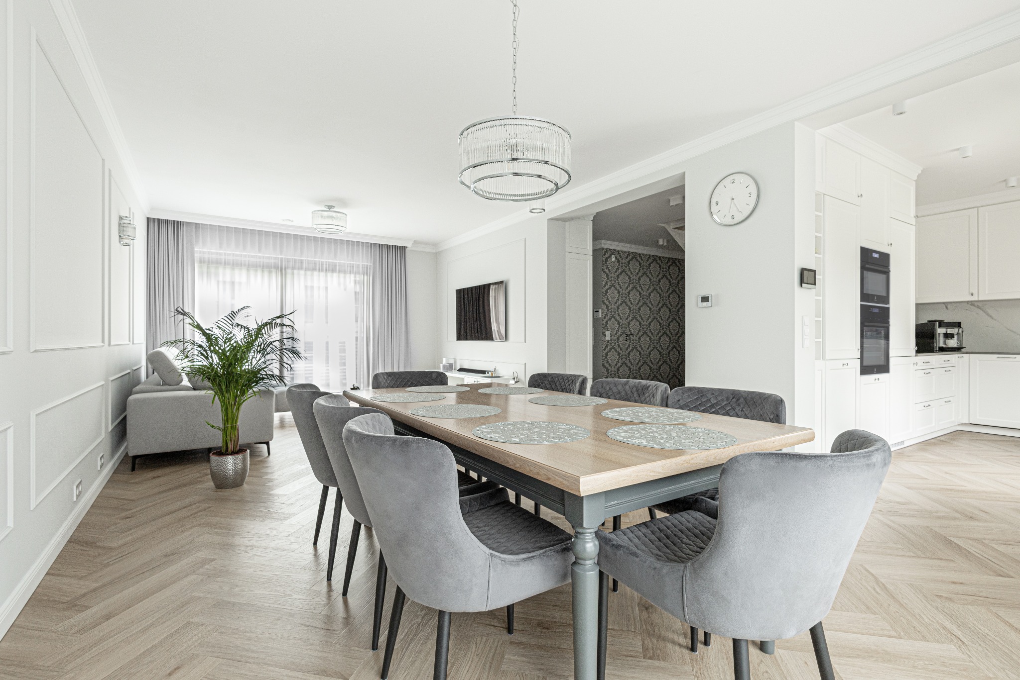 Jasne wnętrze: stół z szarymi krzesłami, parkiet ułożony w jodełkę, widok na salon z szarą sofą i kuchnię z białymi szafkami.