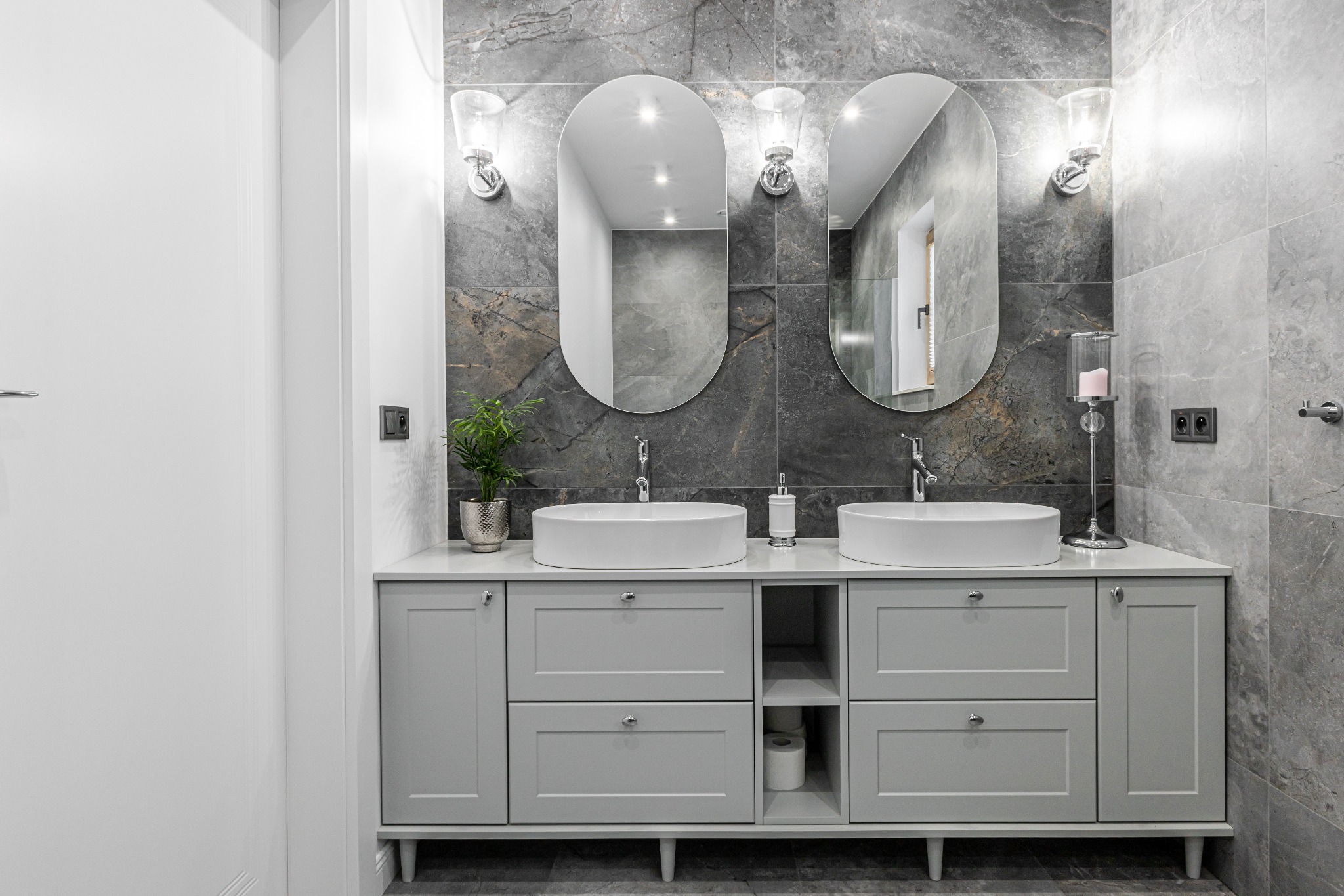 Nowoczesna łazienka z szarą marmurową ścianą, podwójną umywalką nablatową, owalnymi lustrami z oświetleniem i szarą szafką z półkami na zapas papieru toaletowego.