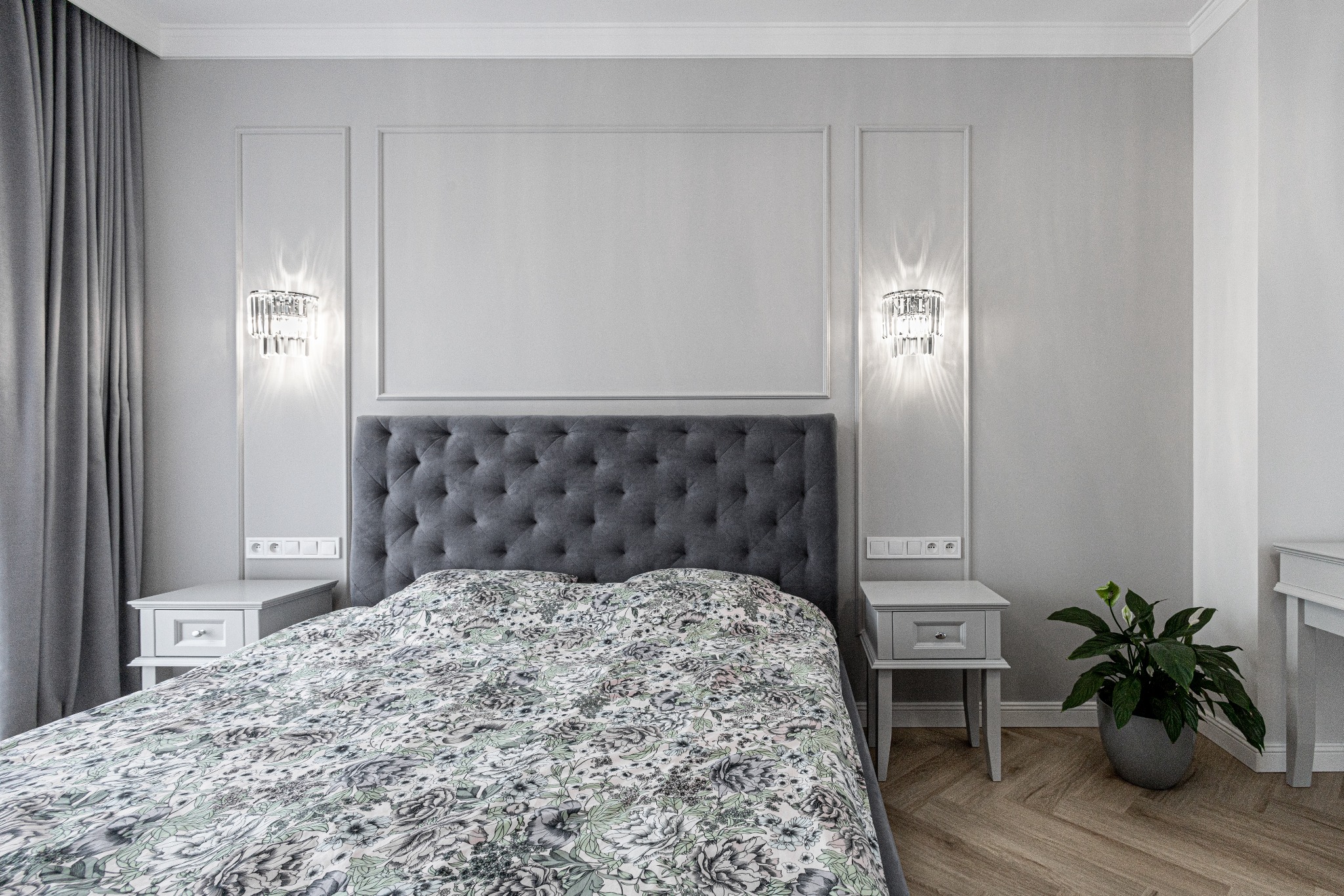 Elegancka sypialnia w odcieniach szarości z podwójnym łóżkiem, pikowanym zagłówkiem, wzorzystą pościelą, kryształowymi kinkietami i parkietem ułożonym w jodełkę.