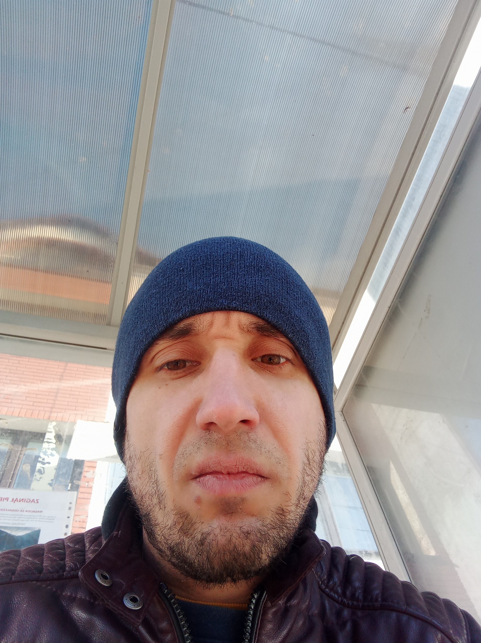 Mężczyzna w czapce stojący pod zadaszeniem z przezroczystych paneli, widoczne elementy konstrukcji i reklama w tle.