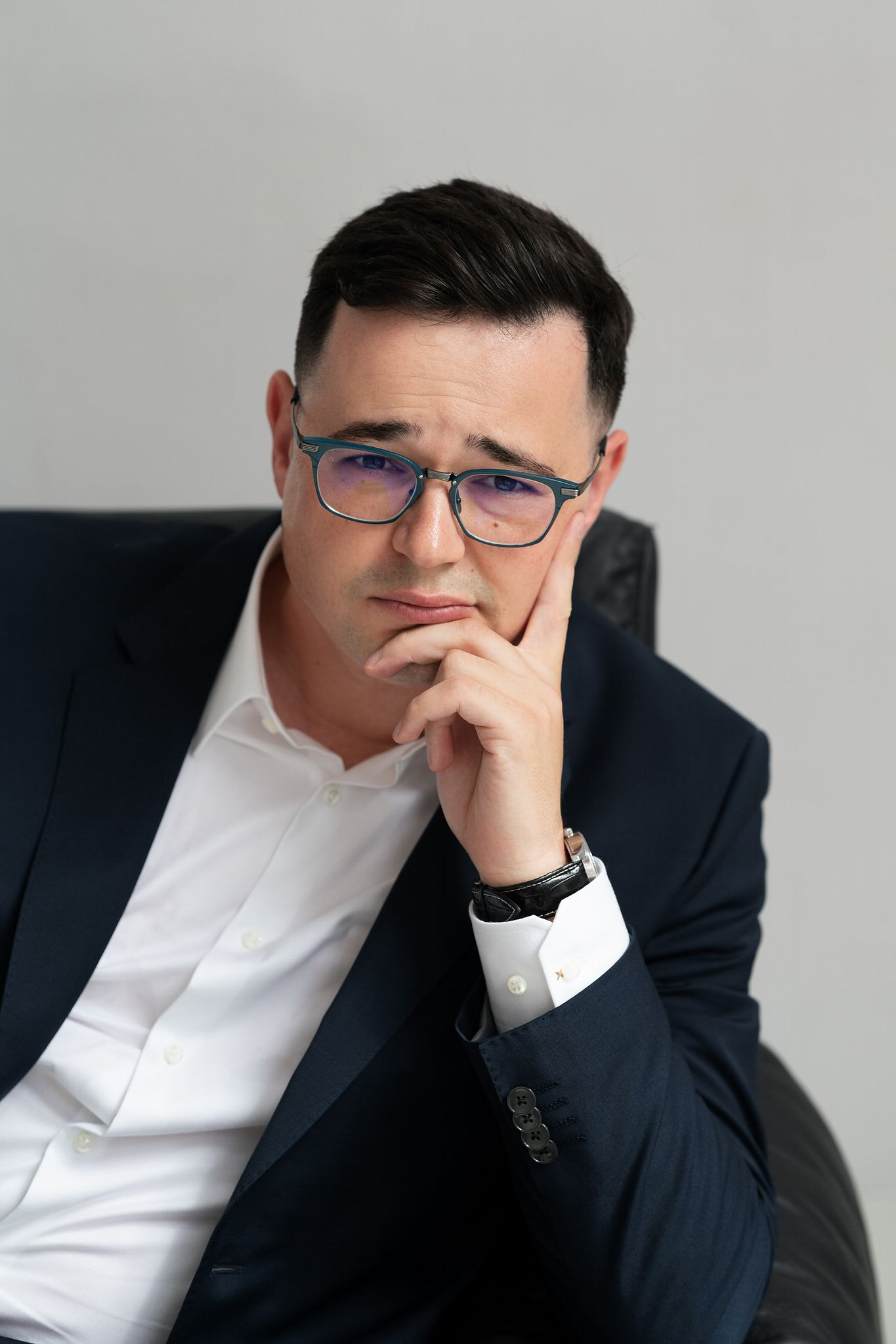 Portret mężczyzny w okularach i granatowym garniturze, zamyślony, opierający brodę na dłoni, widoczny zegarek na skórzanym pasku.