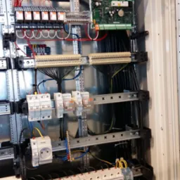 Wnętrze szafy sterowniczej z licznymi przewodami, bezpiecznikami, listwami zaciskowymi i modułem sterującym, zamontowanymi na metalowych szynach wewnątrz obudowy.