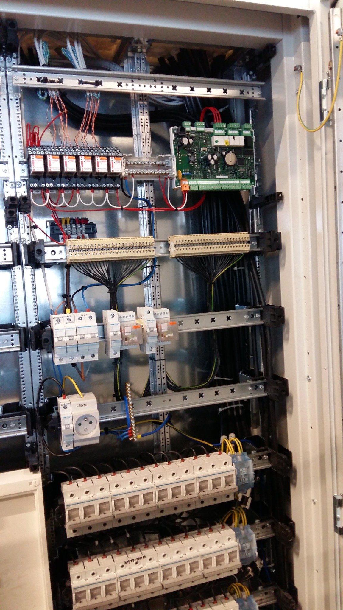 Wnętrze szafy sterowniczej z licznymi przewodami, bezpiecznikami, listwami zaciskowymi i modułem sterującym, zamontowanymi na metalowych szynach wewnątrz obudowy.