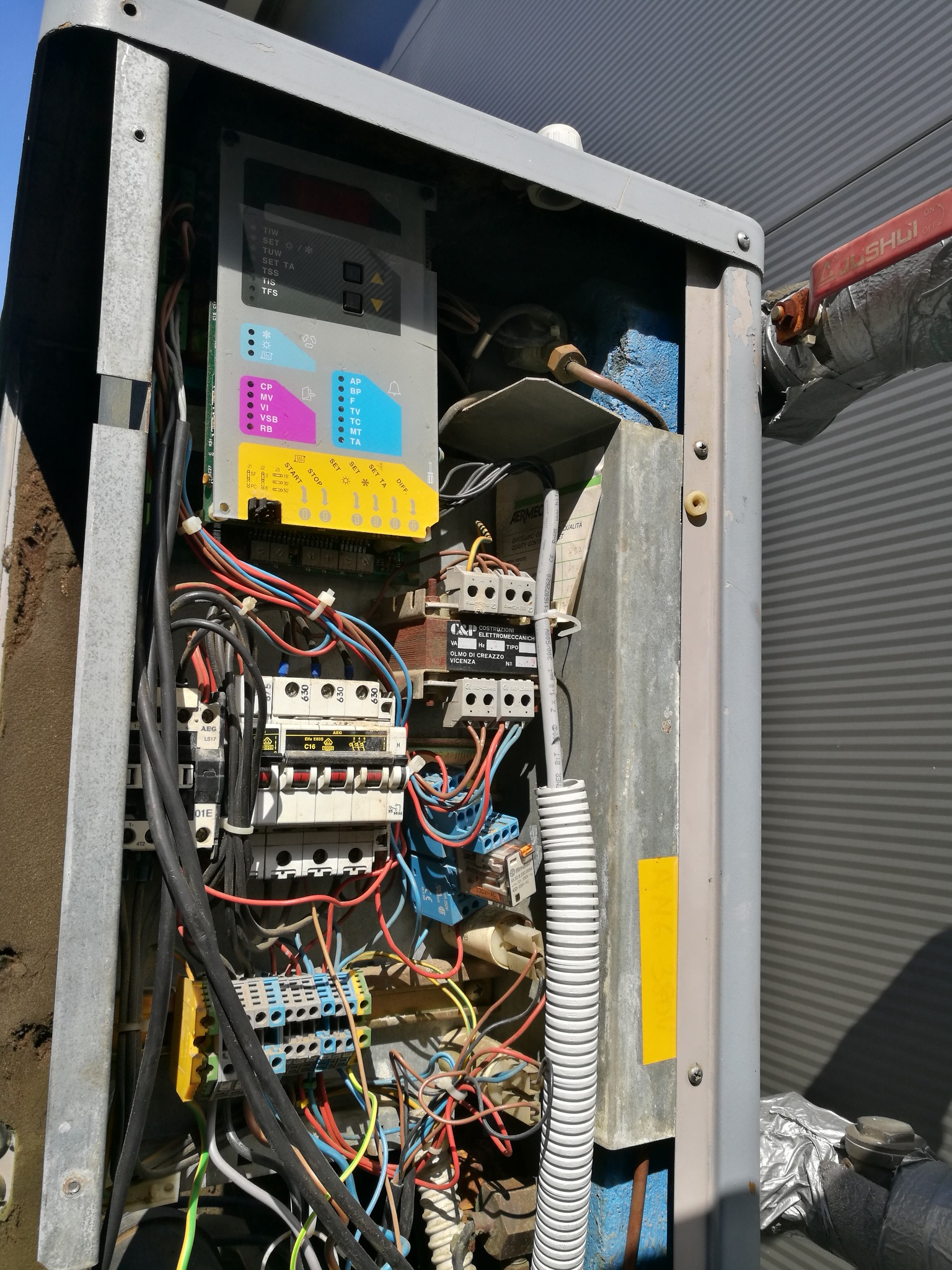 Otwarta skrzynka sterownicza instalacji grzewczej z widocznymi przewodami, bezpiecznikami i panelem kontrolnym z kolorowymi oznaczeniami, rura z zaworem owinięta folią aluminiową po prawej stronie.