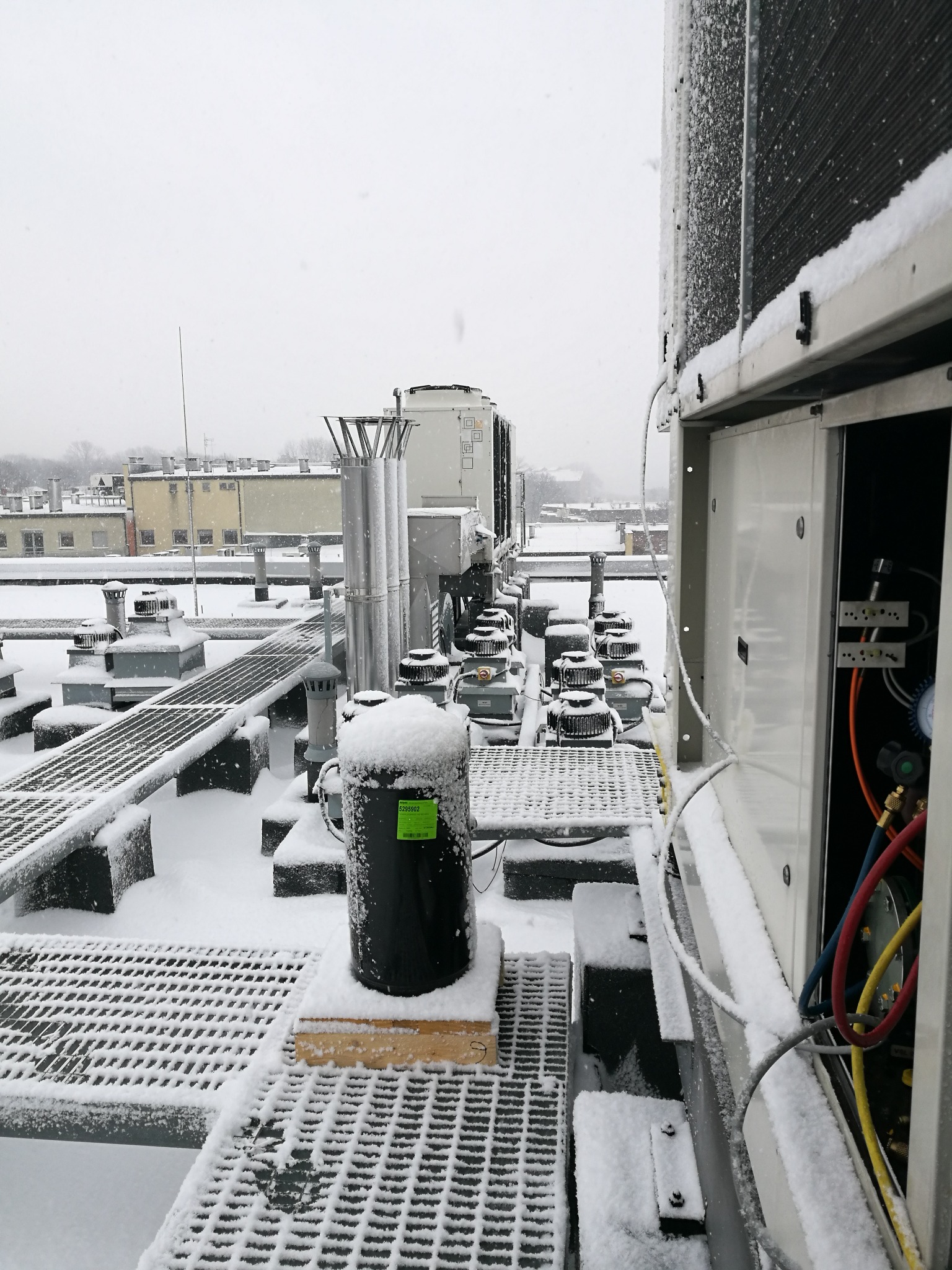 Zimowy krajobraz na dachu budynku z instalacją przemysłową pokrytą śniegiem, widoczne elementy wentylacji, klimatyzacji i rurociągi, otwarte drzwi szafy sterowniczej z przewodami.