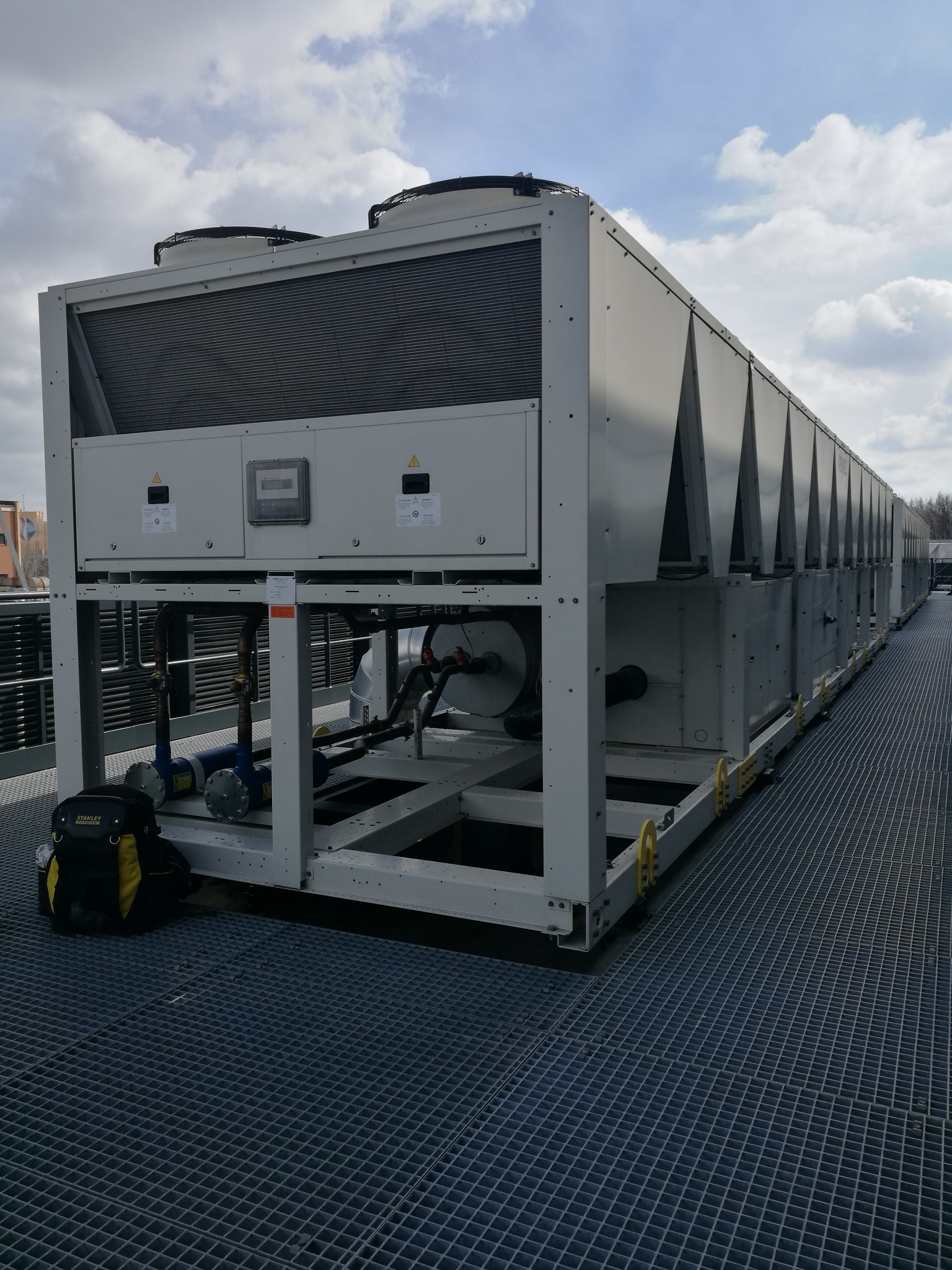 Długa, przemysłowa instalacja chłodnicza z wentylatorami na dachu, widoczna z boku, z torbą narzędziową Stanley na kratownicy pod spodem.